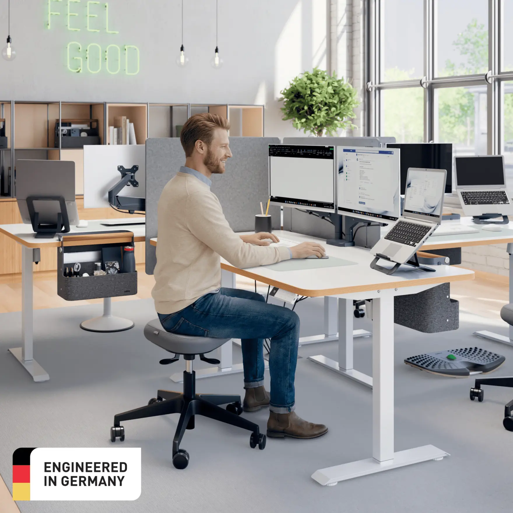 Egy férfi ül egy állítható magasságú asztalnál két monitorral és egy laptoppal.