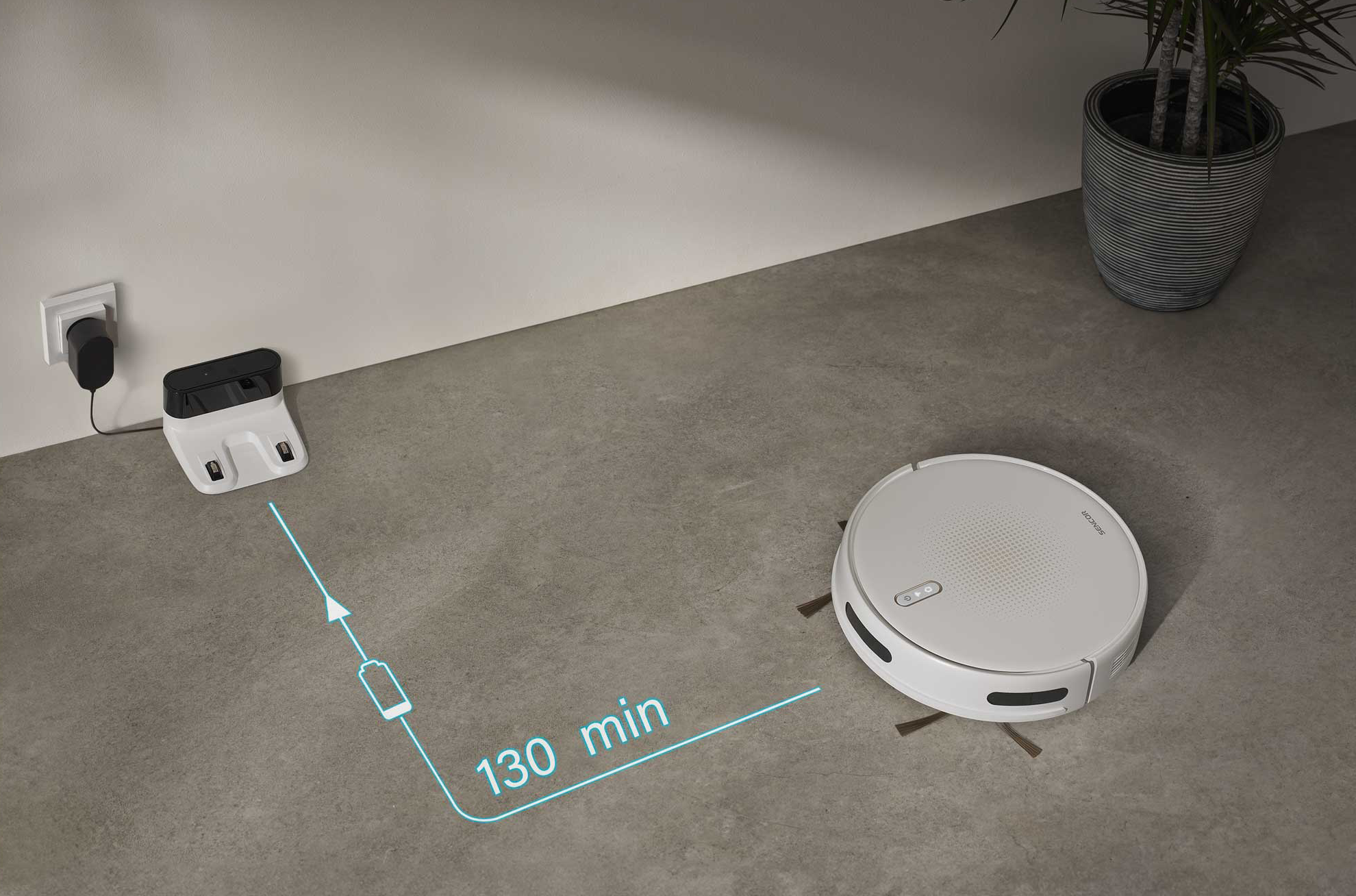 Egy fehér robotporszívó áll egy betonpadlón, egy töltőállomás mellett.