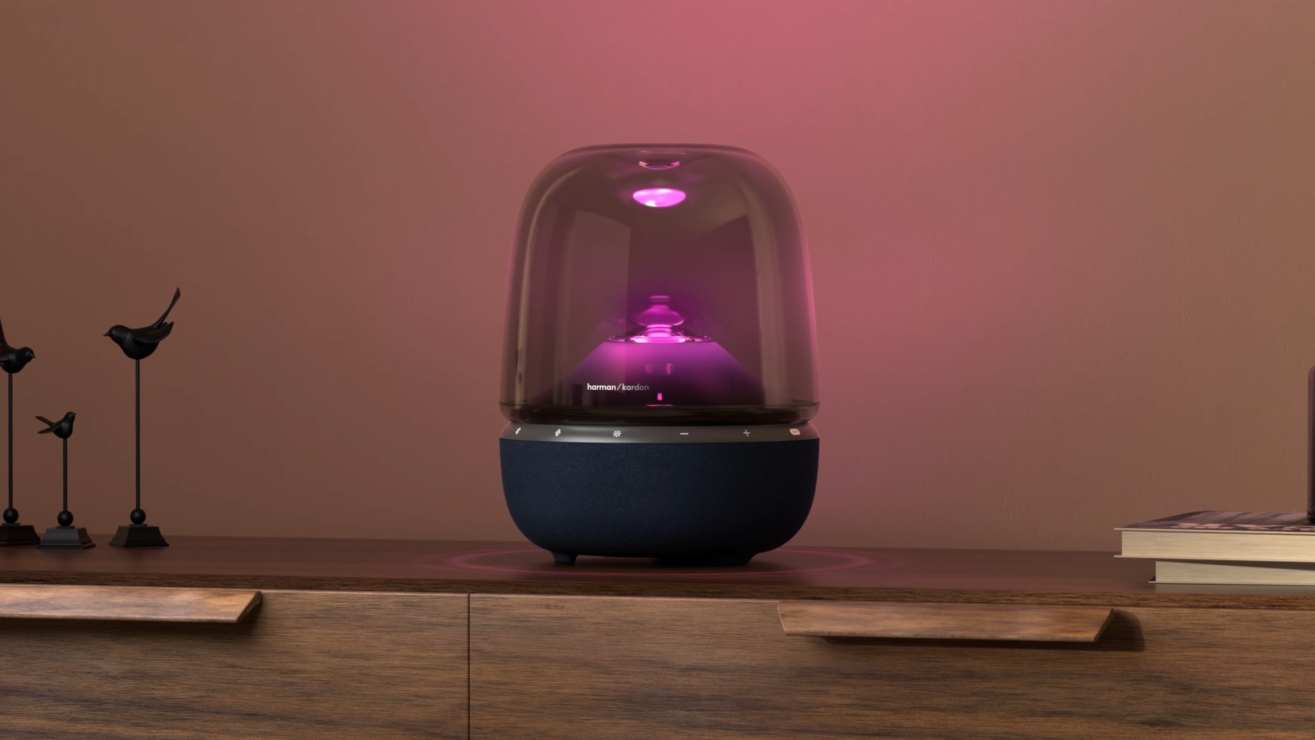 Pembe ışıklı Harman Kardon hoparlör, kitaplar ve kuş heykelleriyle ahşap bir masada duruyor.