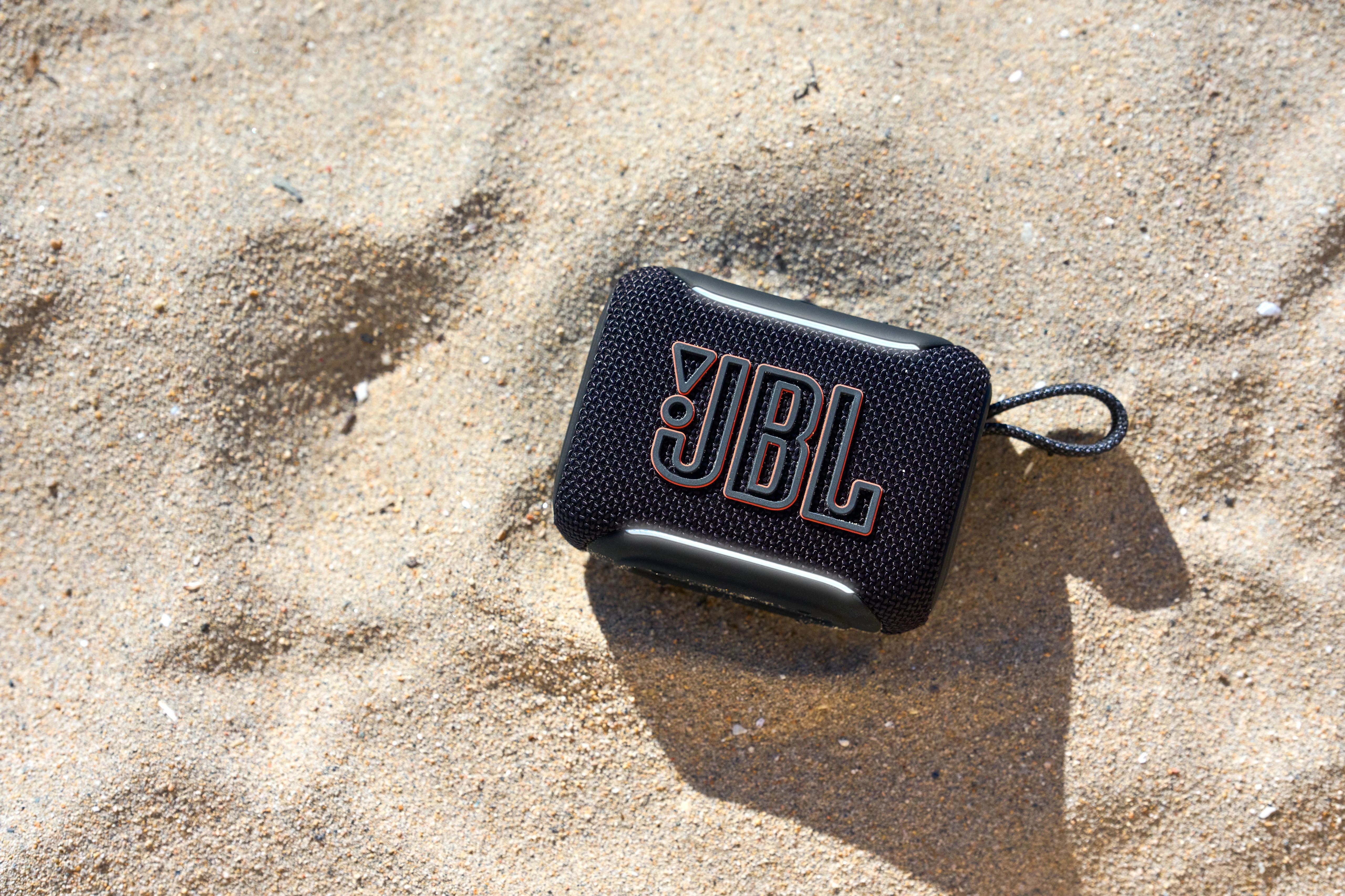 Siyah bir JBL hoparlör kumlu bir plajda duruyor.