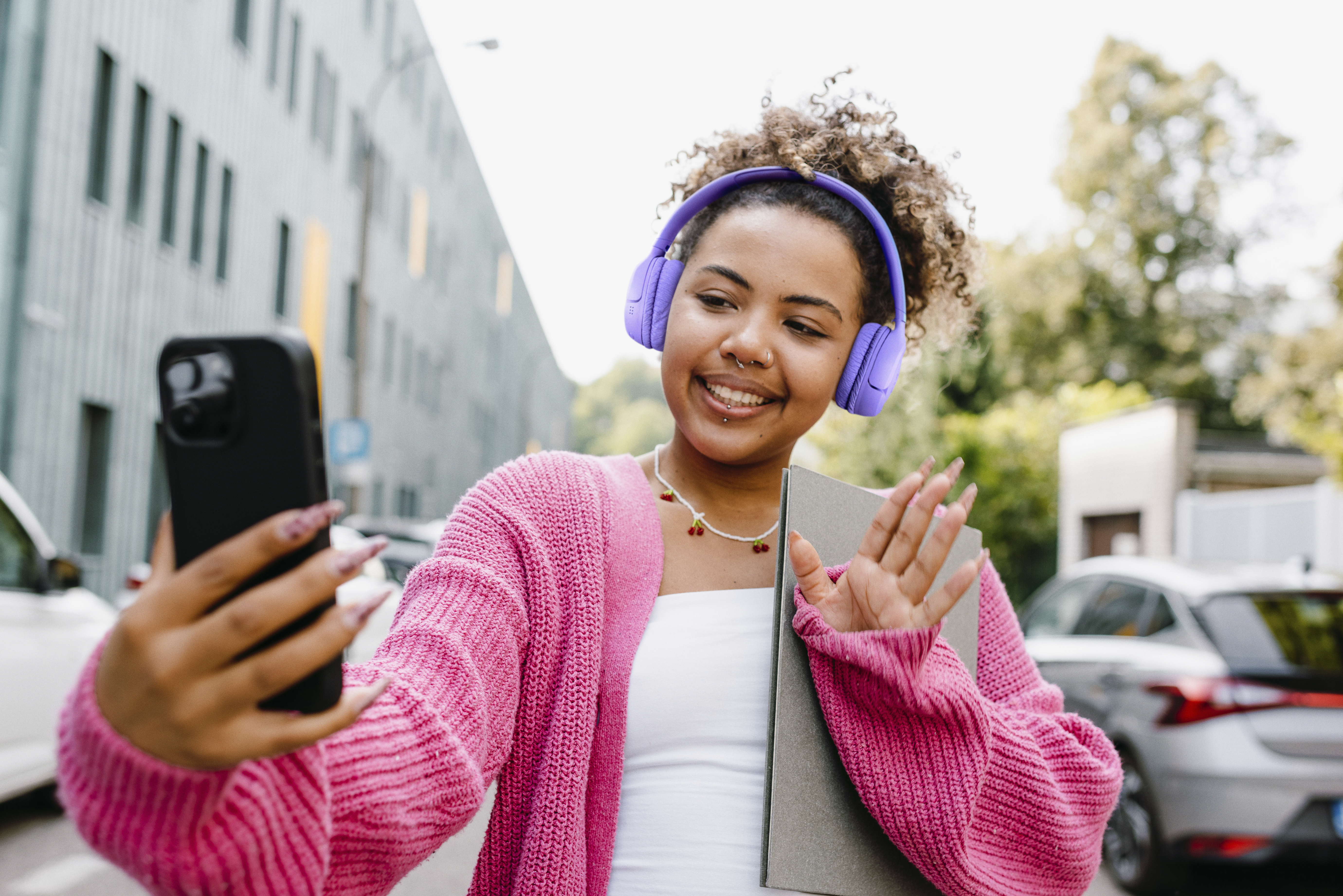 Kulaklıklı gen kadın telefonuyla selfie ekiyor, el sallıyor. Pembe hl giyiyor.