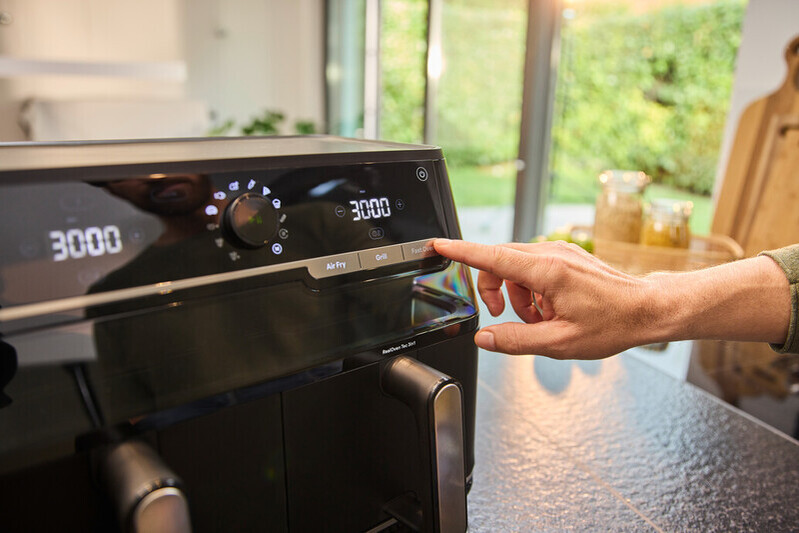 Une main appuie sur le bouton "Fast Cook" d'une friteuse à air noire avec écran numérique.