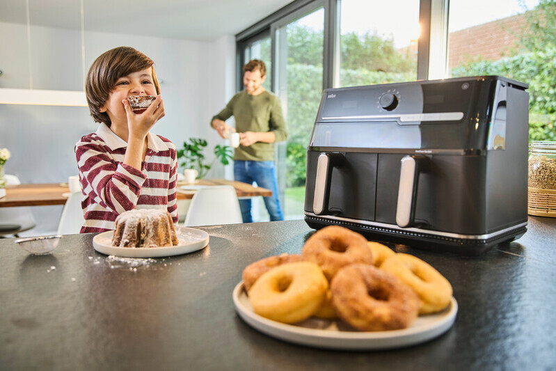 Garçon mange un gâteau, homme à l'arrière-plan, friteuse à air et donuts sur le comptoir.