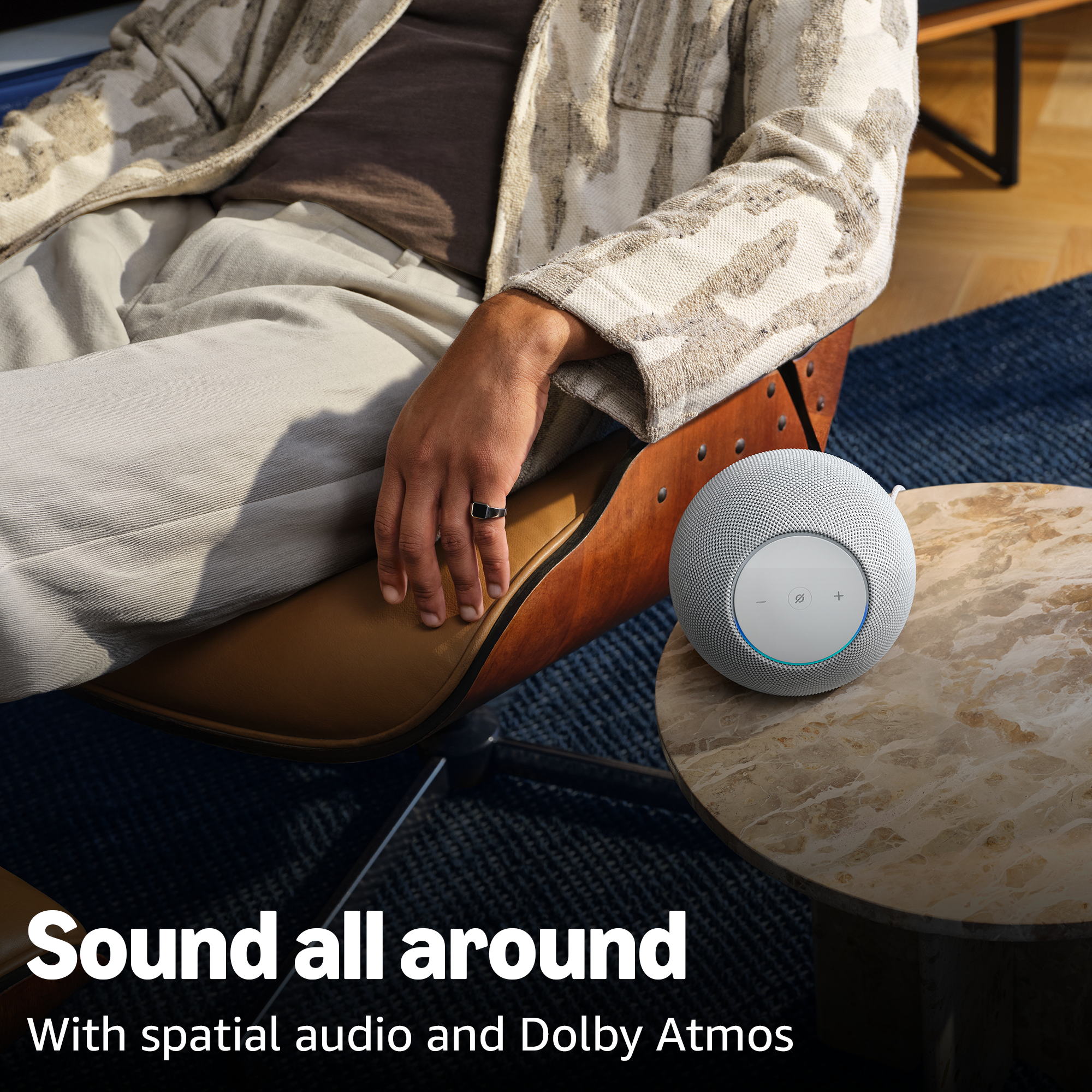 Eine Person sitzt in einem Stuhl neben einem runden Lautsprecher auf einem Tisch. Text: „Rundum-Sound“.