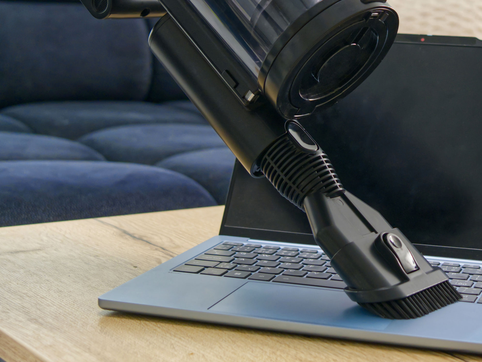 Egy fekete porszívófej tisztít egy kék laptop billentyűzetet egy faasztalon.