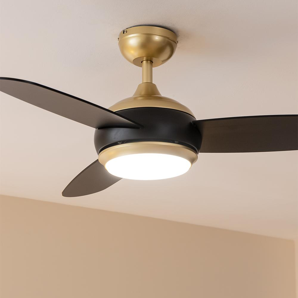 Un ventilador de techo moderno con aspas negras y cuerpo de latón y negro, iluminado por una luz central.