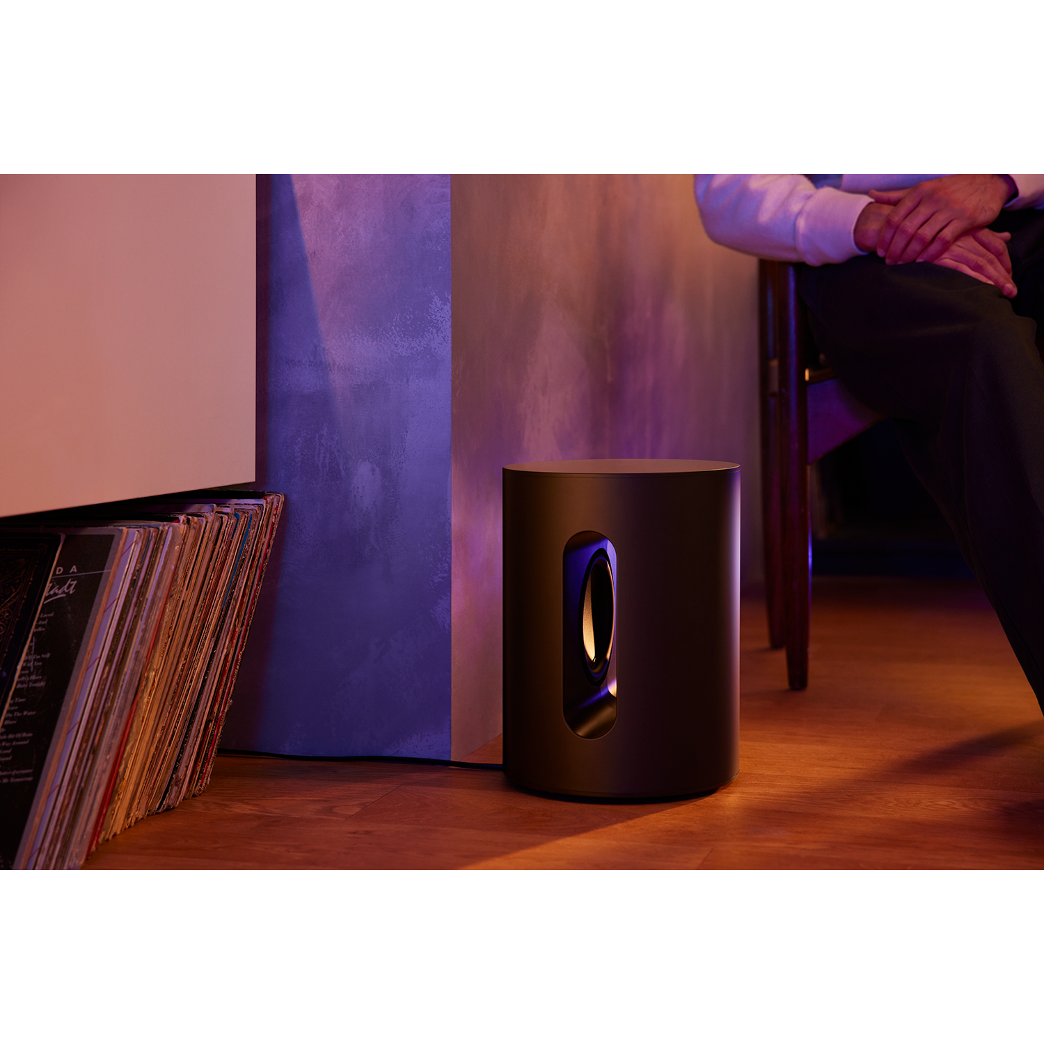 Een zwarte subwoofer op houten vloer naast platen. Een persoon zit op de achtergrond.