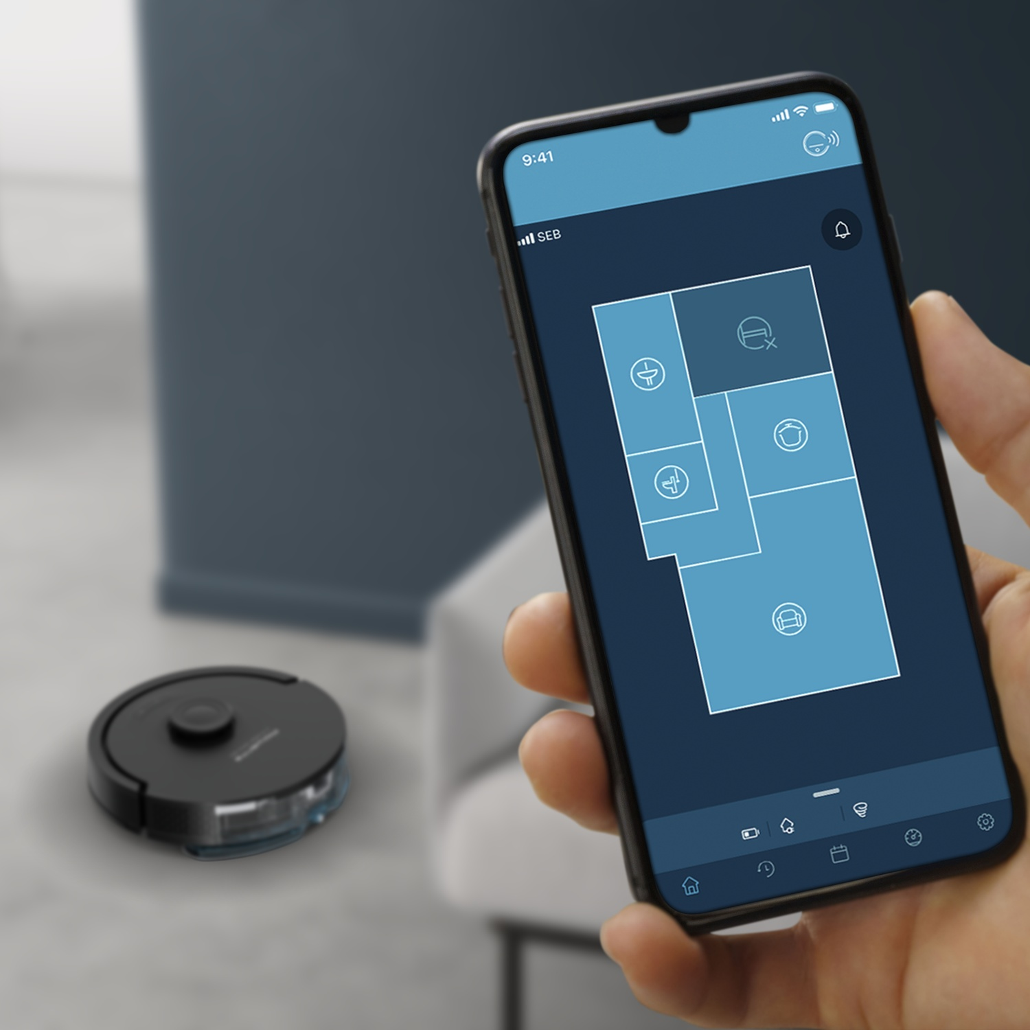 A smartphone shows a room map of a robot vacuum. A robot vacuum is on the floor.