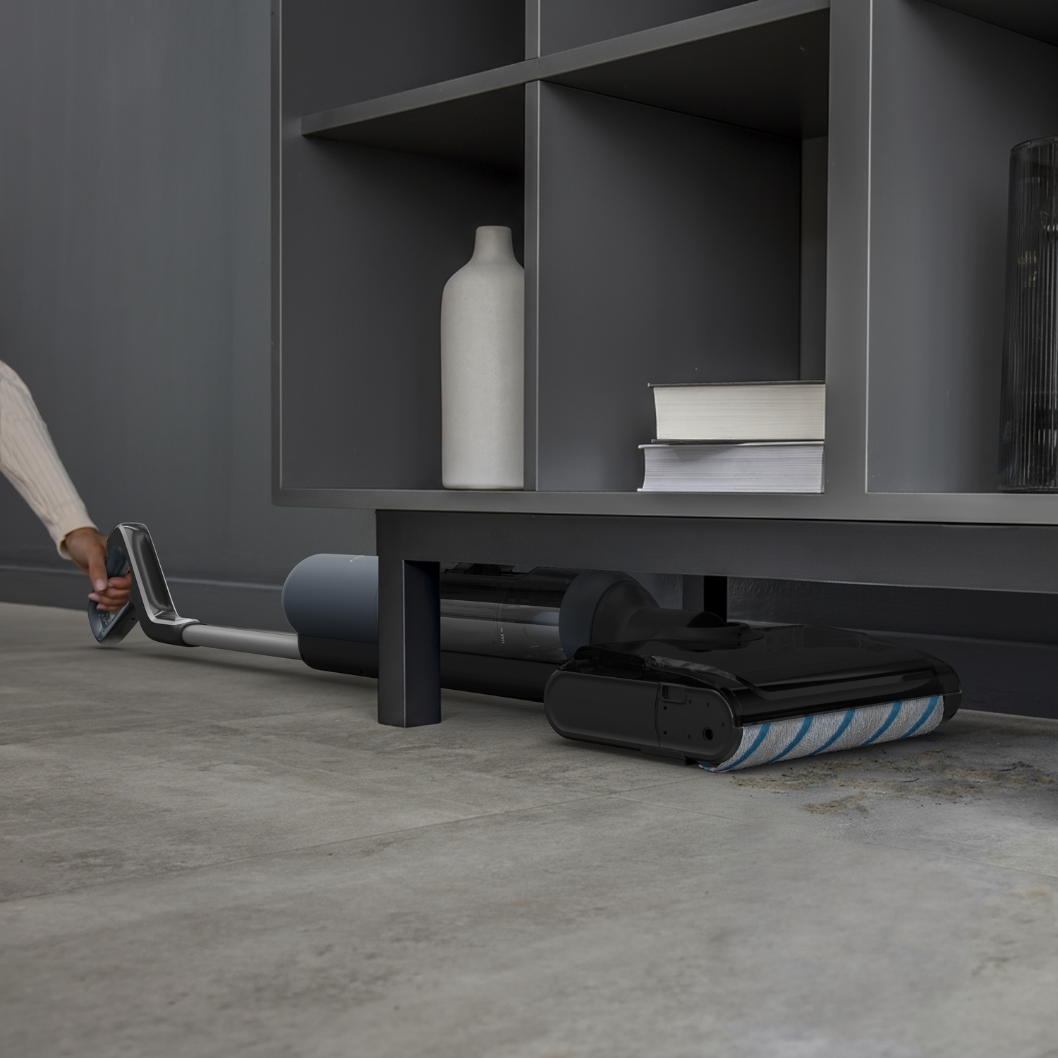 A hand uses a vacuum cleaner under a shelf with books and a vase.