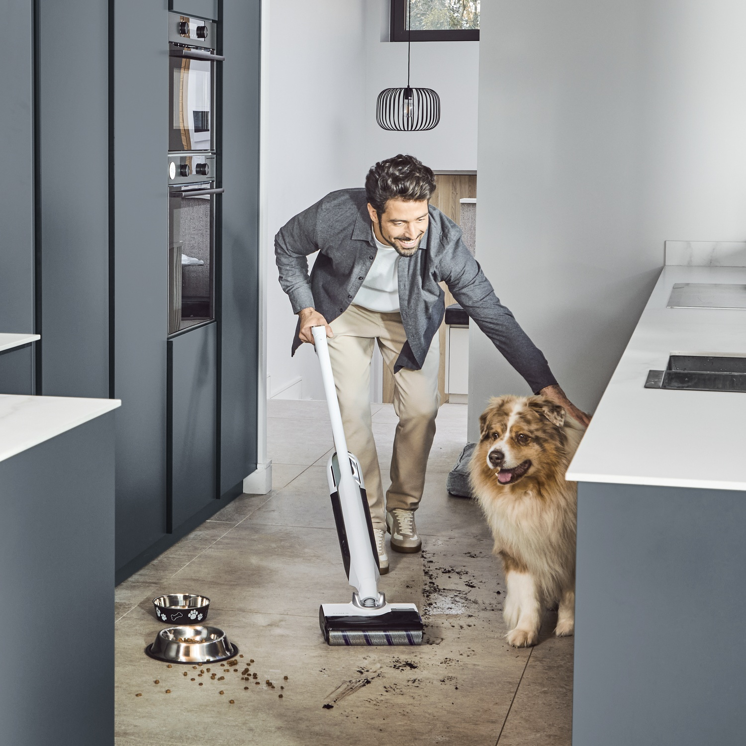 Man stofzuigt de vloer terwijl een hond toekijkt. Twee voerbakken staan ernaast.