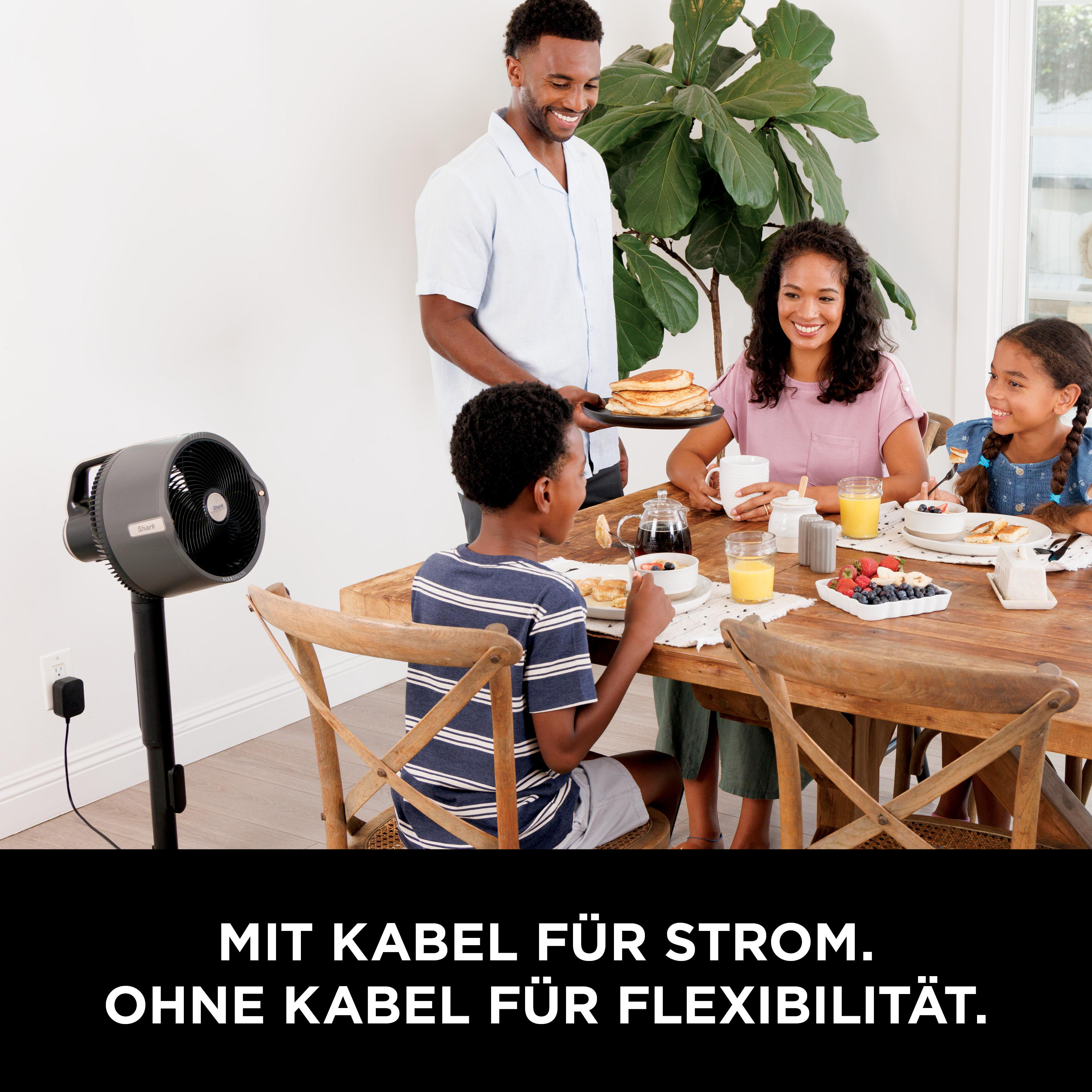 Familie isst Frühstück am Holztisch, Mann serviert Pfannkuchen, Ventilator im Hintergrund.