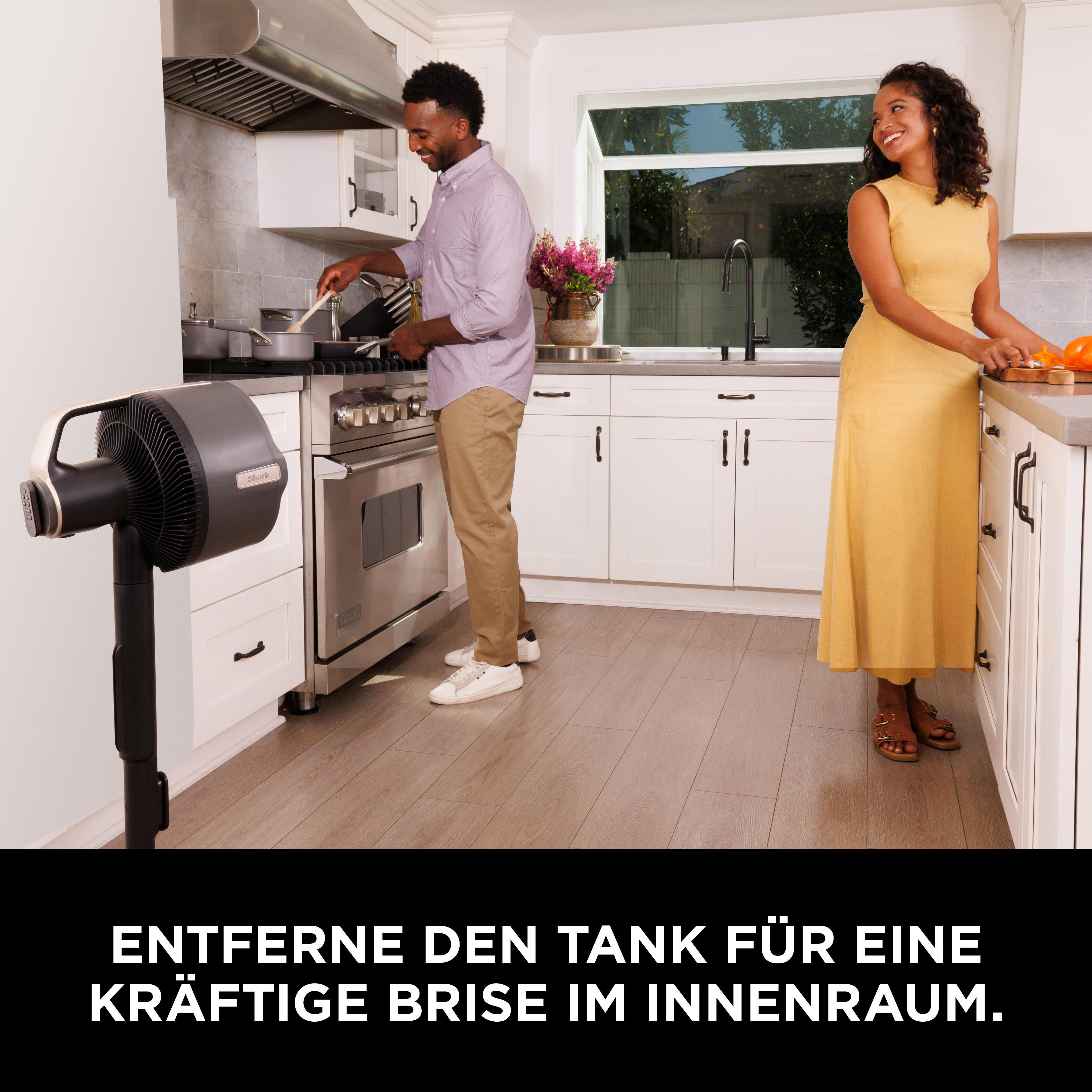 Mann kocht, Frau steht am Tresen. Ein Ventilator steht im Vordergrund.