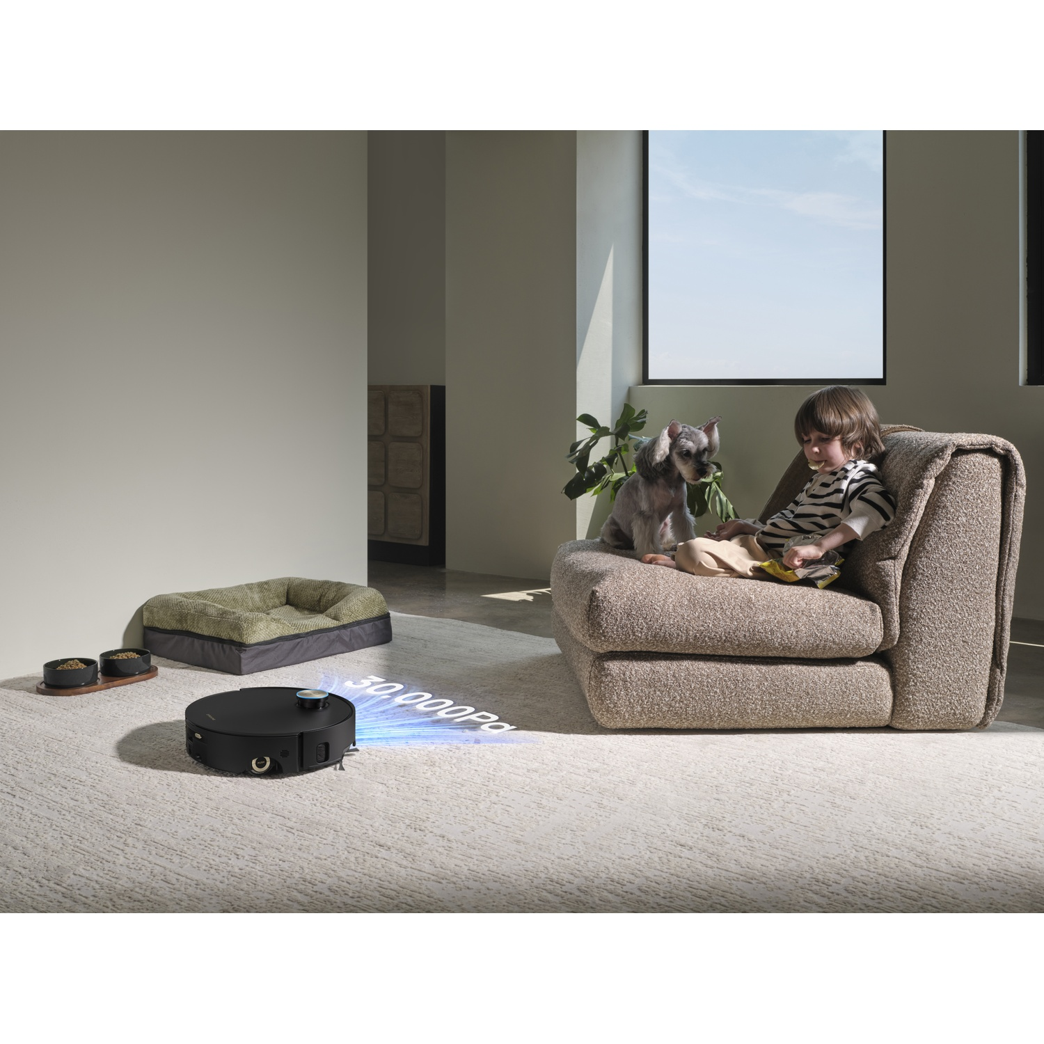 A robot vacuum cleans a carpet while a boy and a dog sit on a sofa.