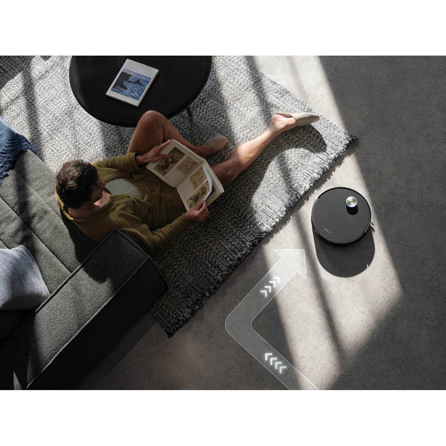 A man reads on a sofa while a robot vacuum cleans the floor.