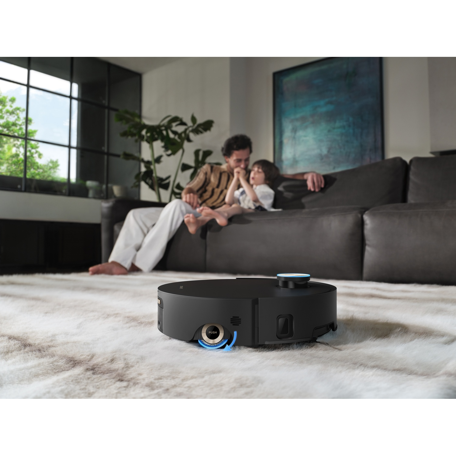 A black robot vacuum cleans a fluffy rug while a man and child relax on a sofa.