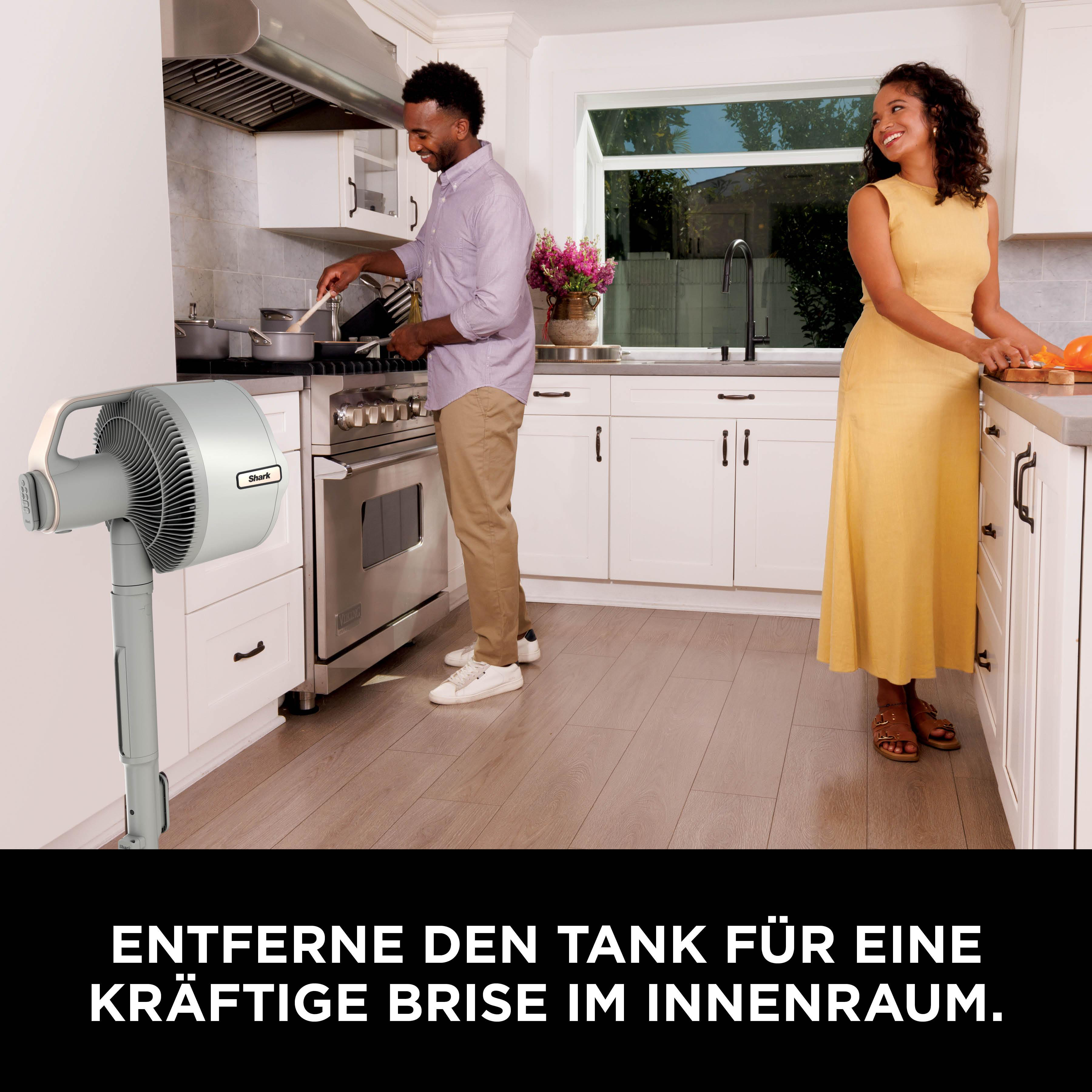 Ein Mann kocht, eine Frau schneidet Obst in einer hellen Küche. Ein Ventilator steht im Vordergrund.