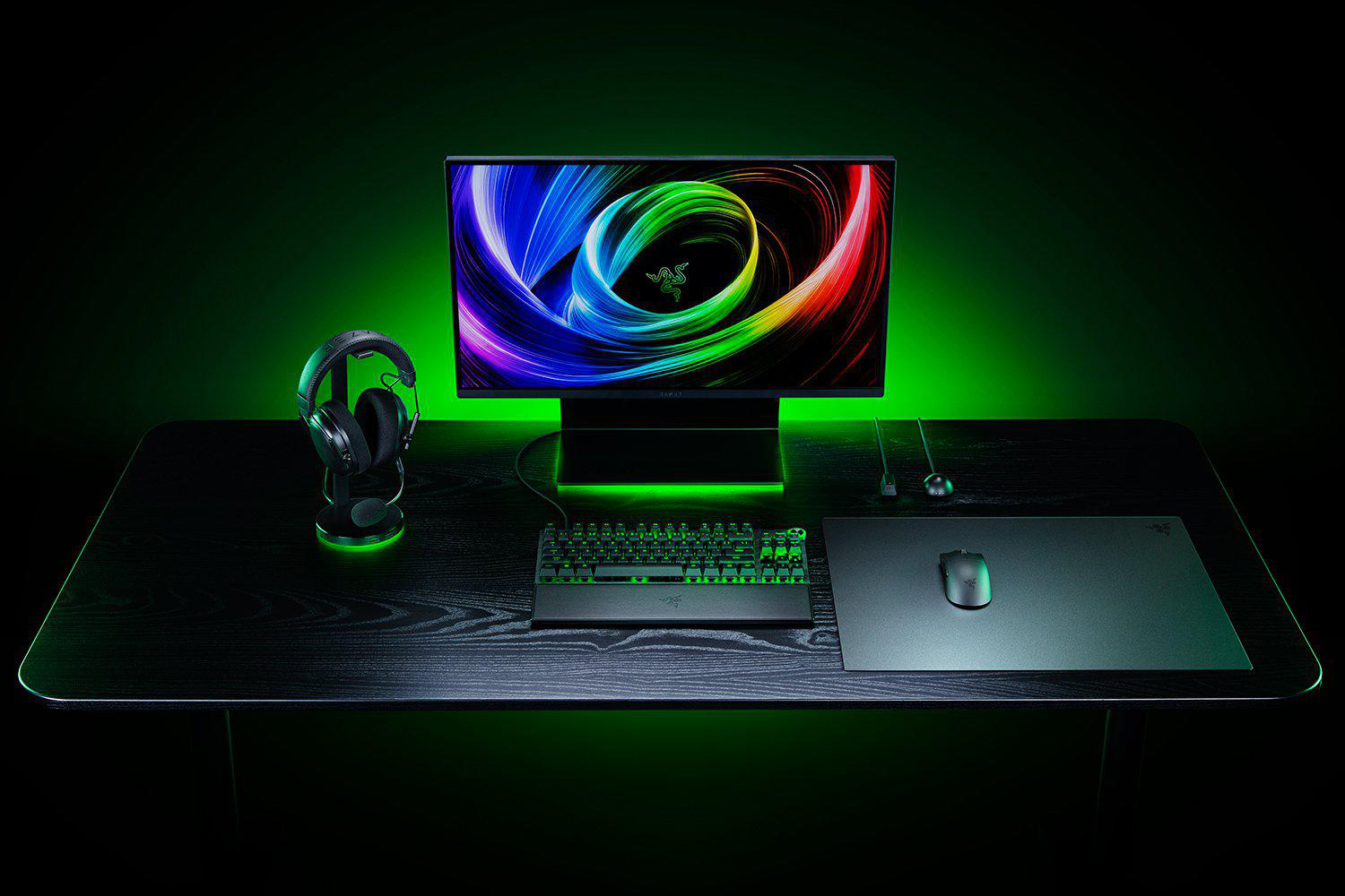 Ein schwarzer Gaming-Schreibtisch mit Monitor, Tastatur, Maus und Kopfhörern. Grüne Umgebungsbeleuchtung.