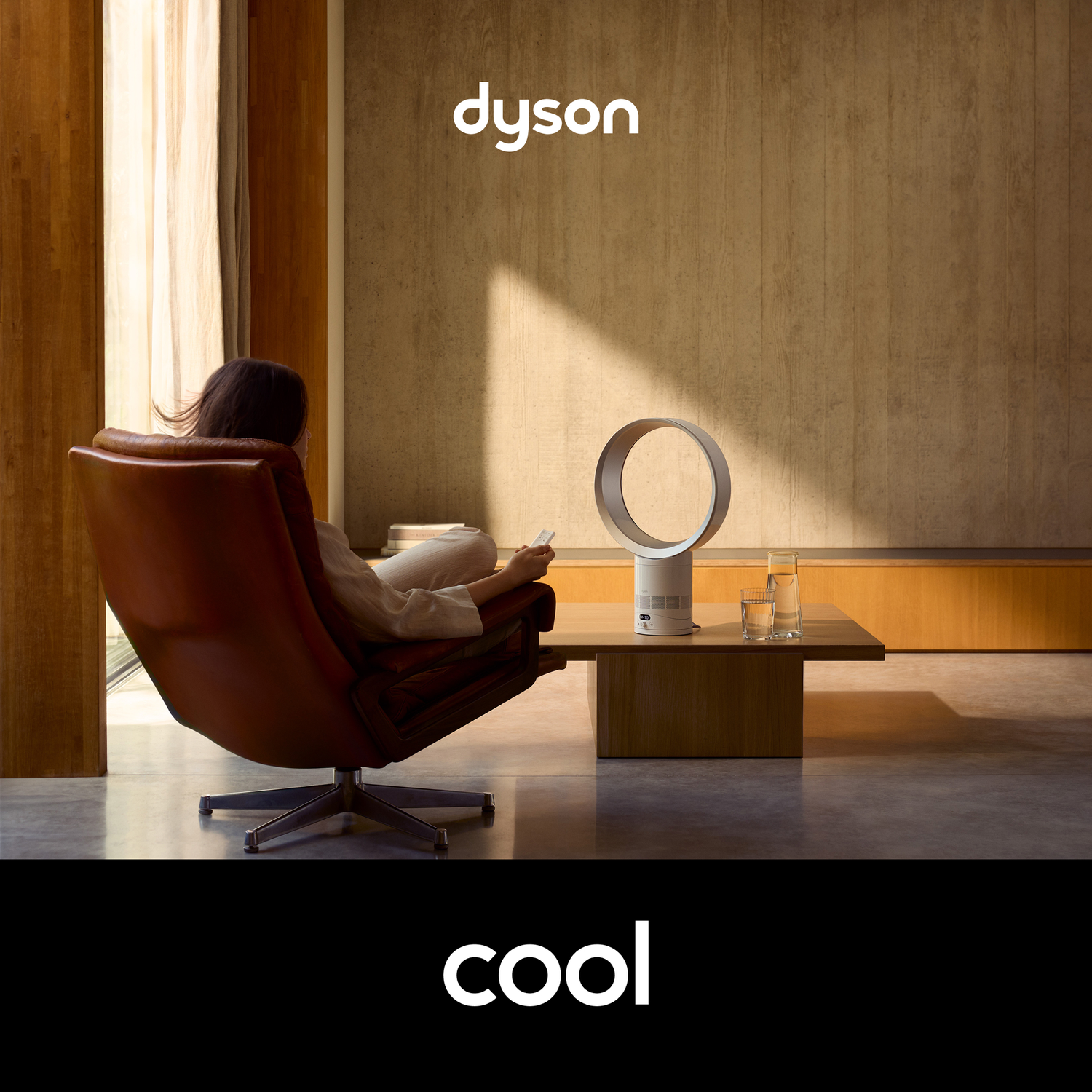 A woman sits in a brown leather chair, facing a Dyson fan and two glasses of water on a wooden table. Sunlight streams in from a window.