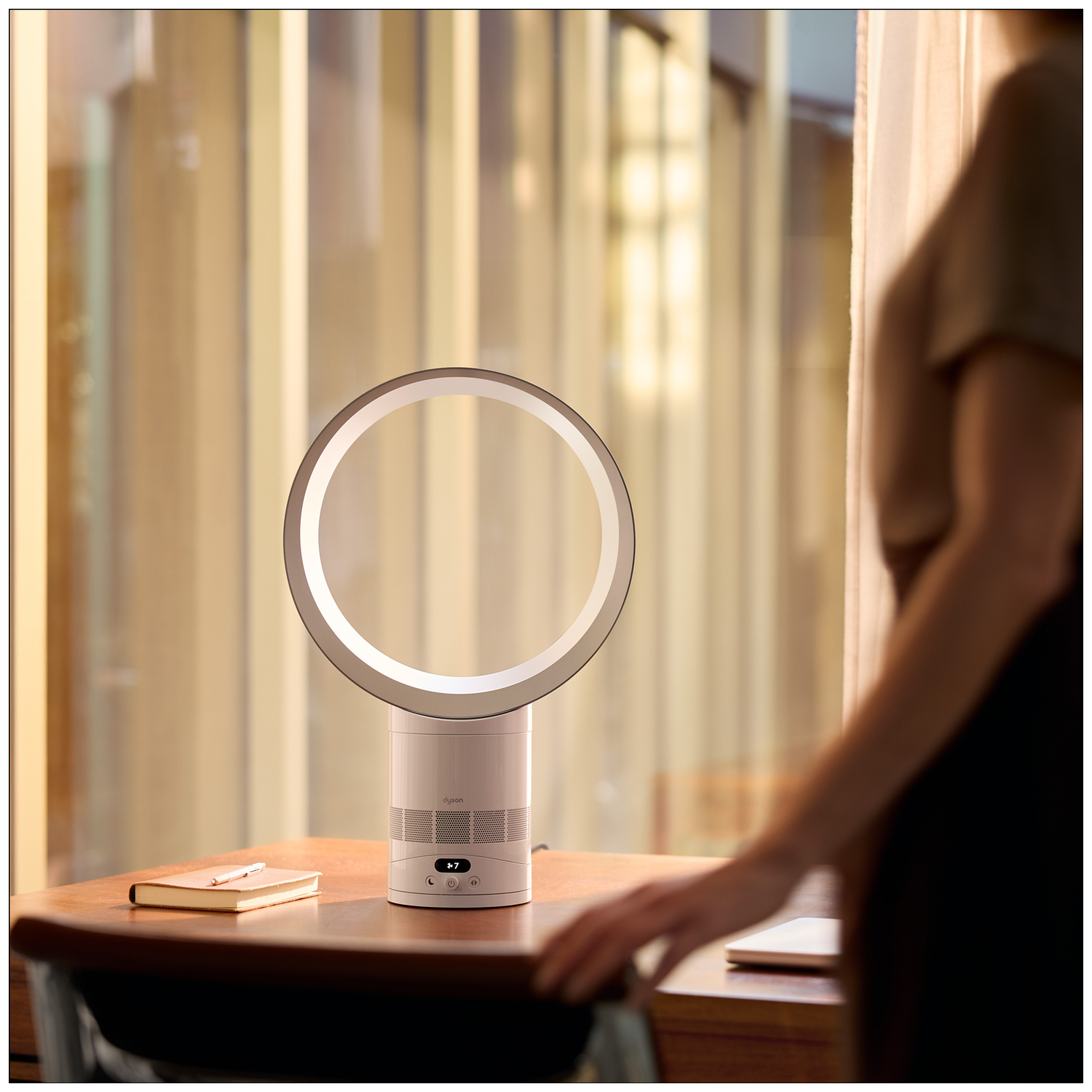 A white bladeless fan sits on a wooden desk next to a notebook and pen. A person stands to the right.