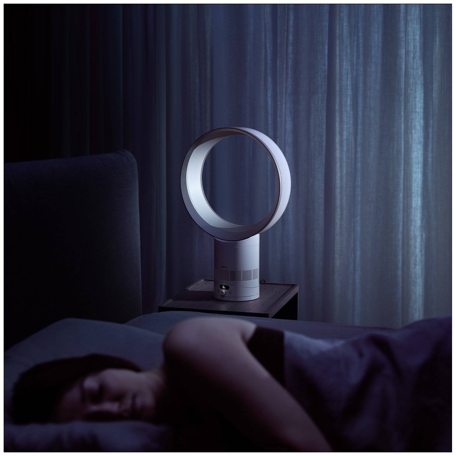 A person sleeps in bed, a ring fan stands on the nightstand.