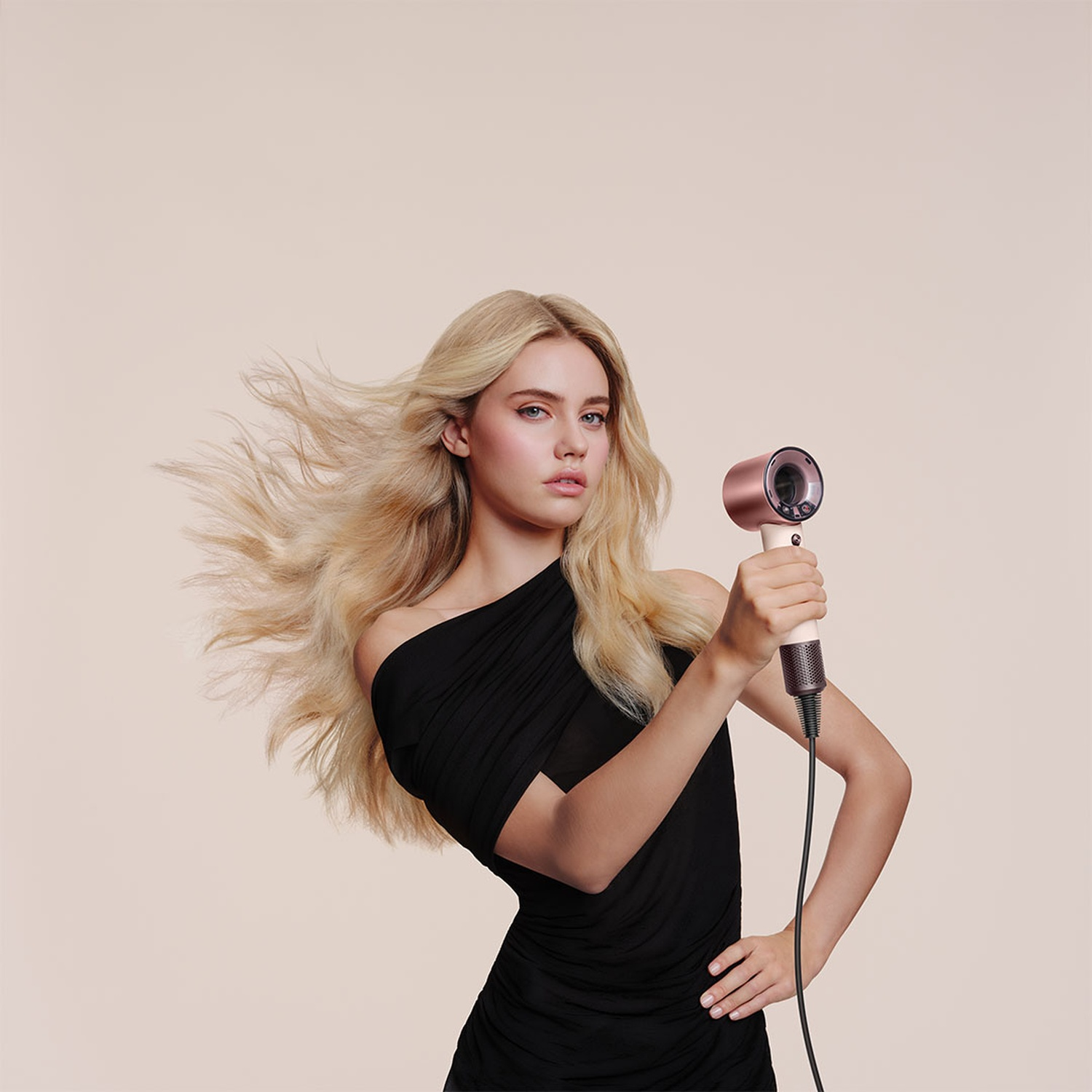 A blonde woman in a black dress holds a rose gold hairdryer with a white handle.