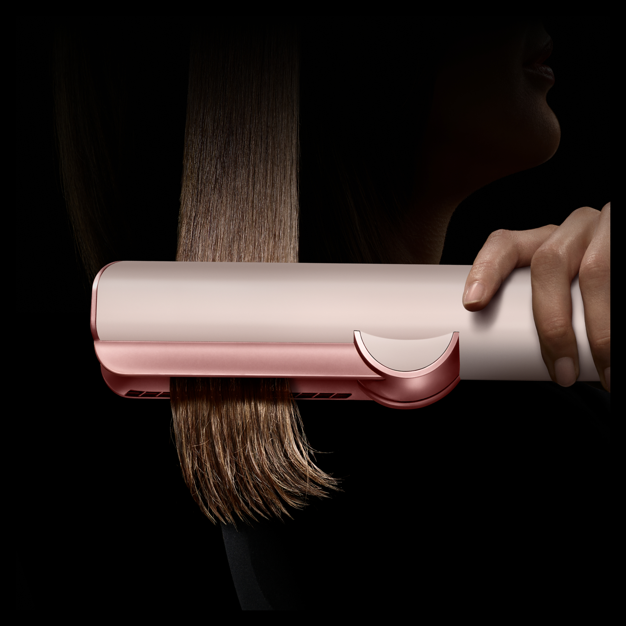 Eine Hand benutzt einen roségoldenen Haarglätter an braunem Haar vor schwarzem Hintergrund.