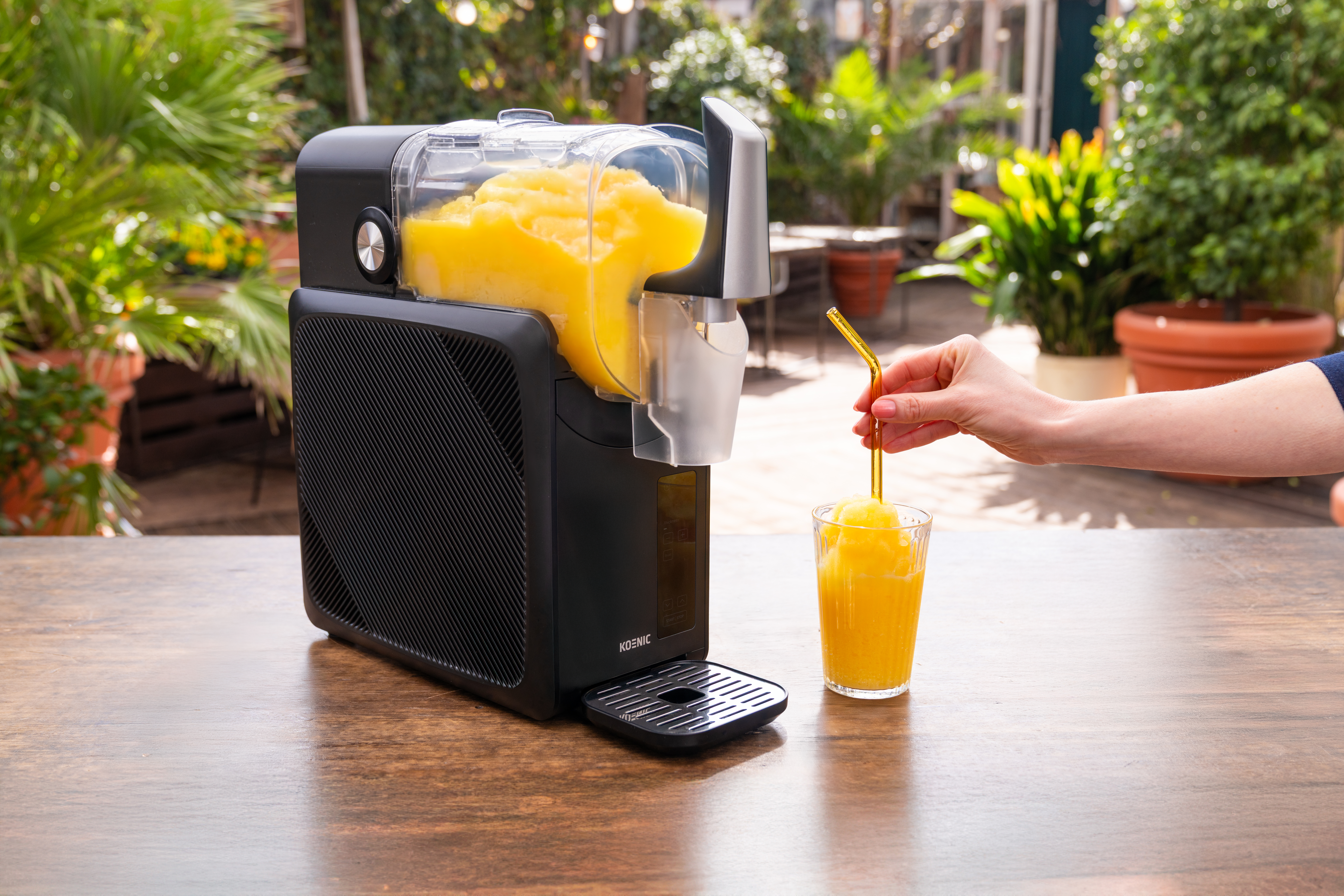 Een zwarte slushmachine met geel bevroren drankje, ernaast een glas met drankje en rietje.