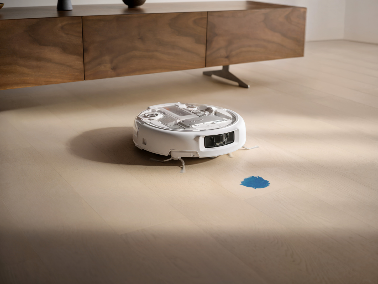 Un robot aspirateur blanc nettoie un déversement bleu sur un plancher en bois clair.