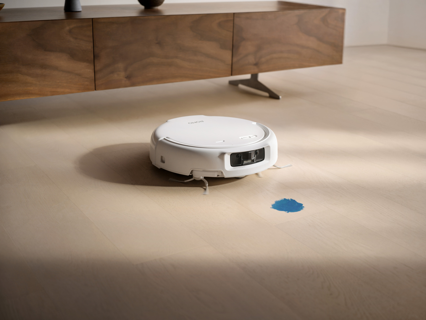 Un robot aspirapolvere bianco pulisce un pavimento in legno accanto a una macchia blu.