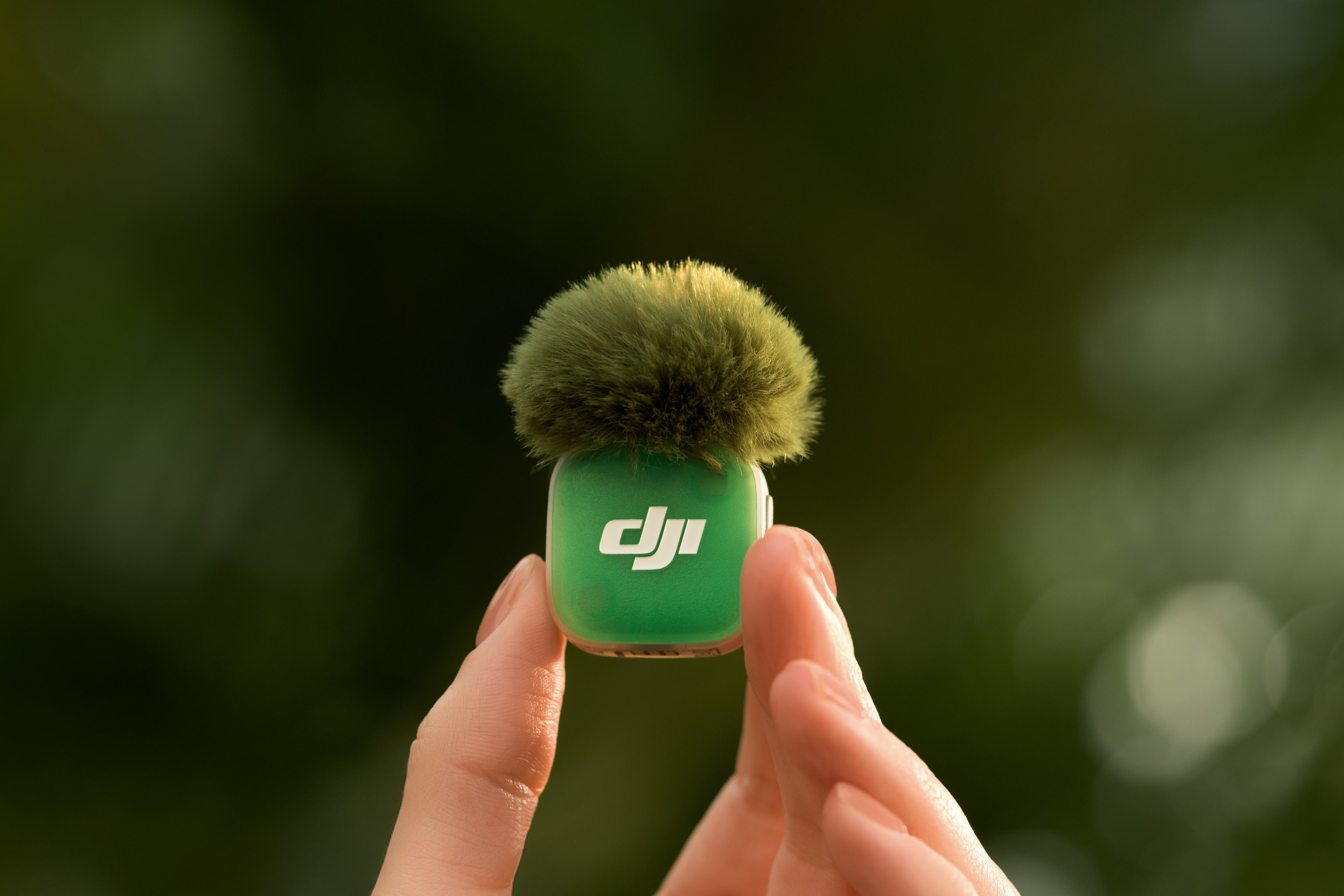 Microphone DJI vert avec dessus poilu, tenu par des doigts sur fond flou.