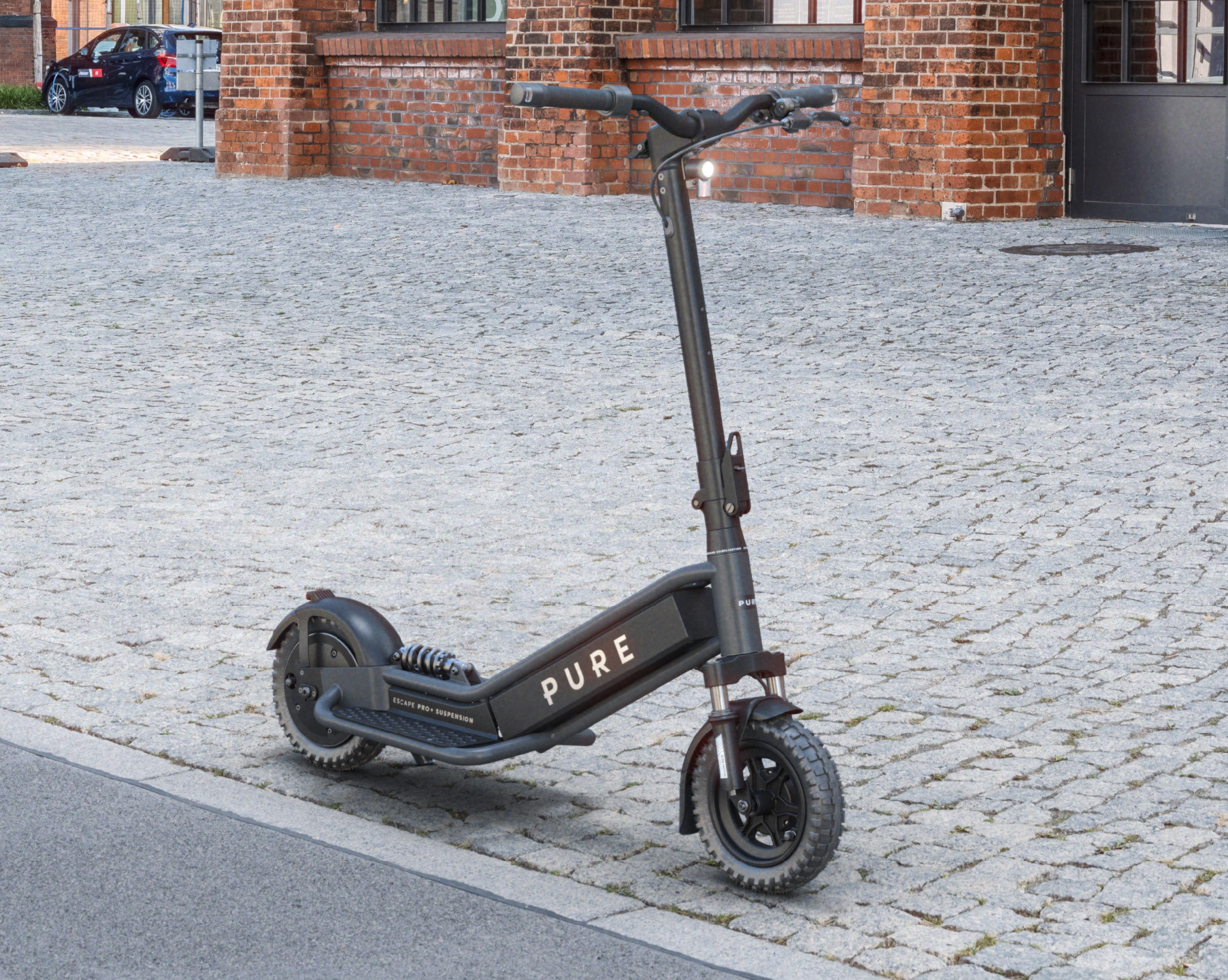 Schwarzer E-Scooter mit Federung und Licht auf Kopfsteinpflaster vor Backsteinhaus.