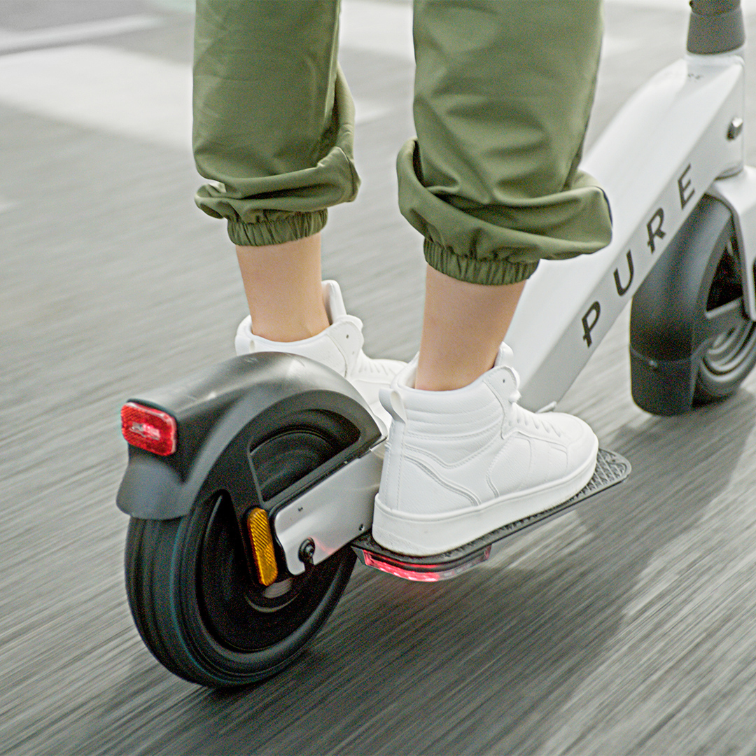 Person in weißen Schuhen und grünen Hosen fährt E-Scooter mit rotem Rücklicht.