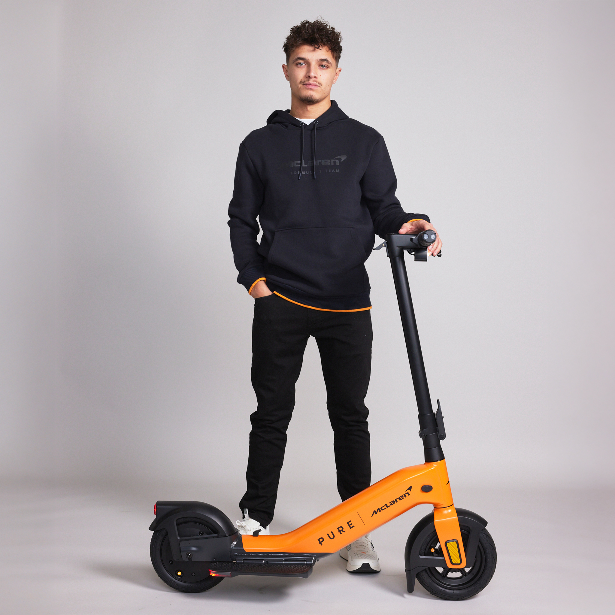 Ein Mann in einem schwarzen Hoodie und Hose steht neben einem orangefarbenen Elektroroller.