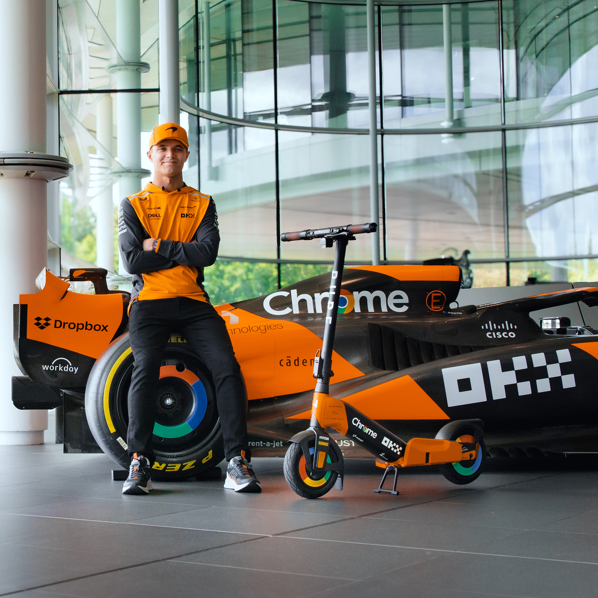 Ein Mann in orangefarbener Kleidung sitzt neben einem Formel-1-Auto und einem Roller.