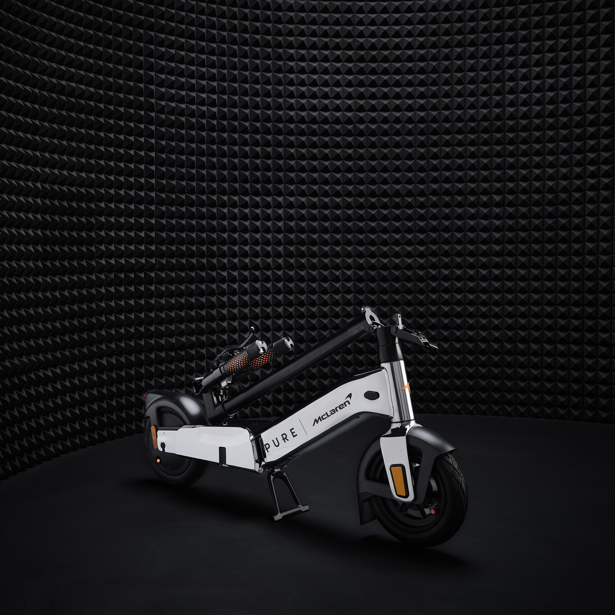 Ein weißer E-Scooter mit McLaren-Logo steht auf schwarzem Untergrund vor schwarzer Wand.
