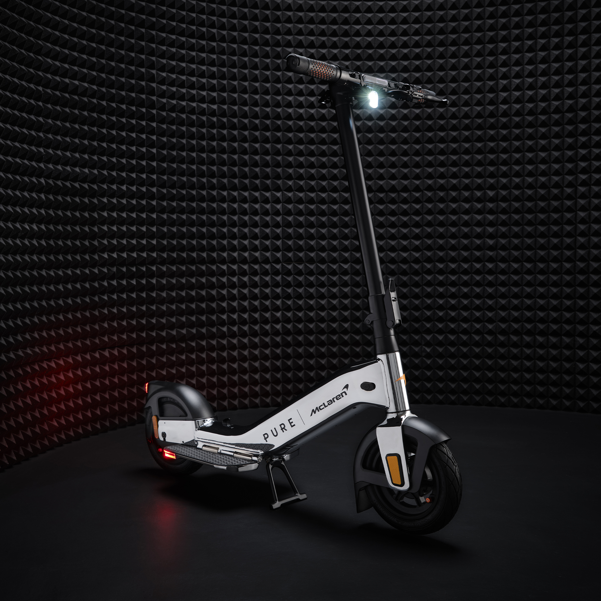 Ein weißer Elektroroller mit McLaren-Logo steht vor einer schwarzen, gemusterten Wand.