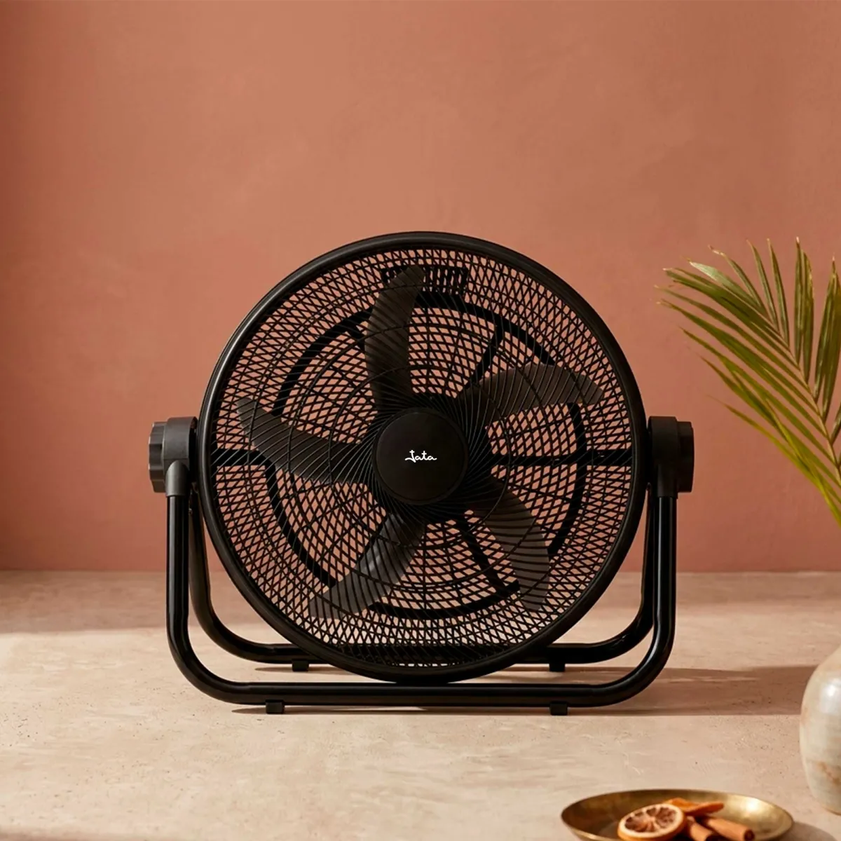 Un ventilador negro se sienta en una mesa con una pared rosada al fondo.