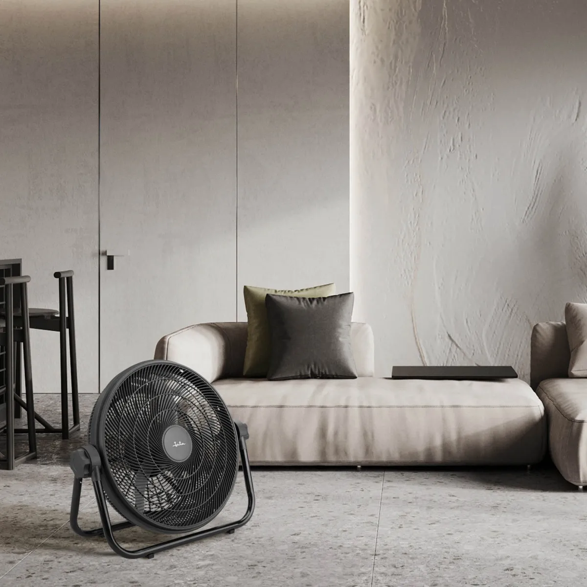 Un ventilador negro se encuentra sobre un suelo gris frente a un sofá marrón claro con cojines.