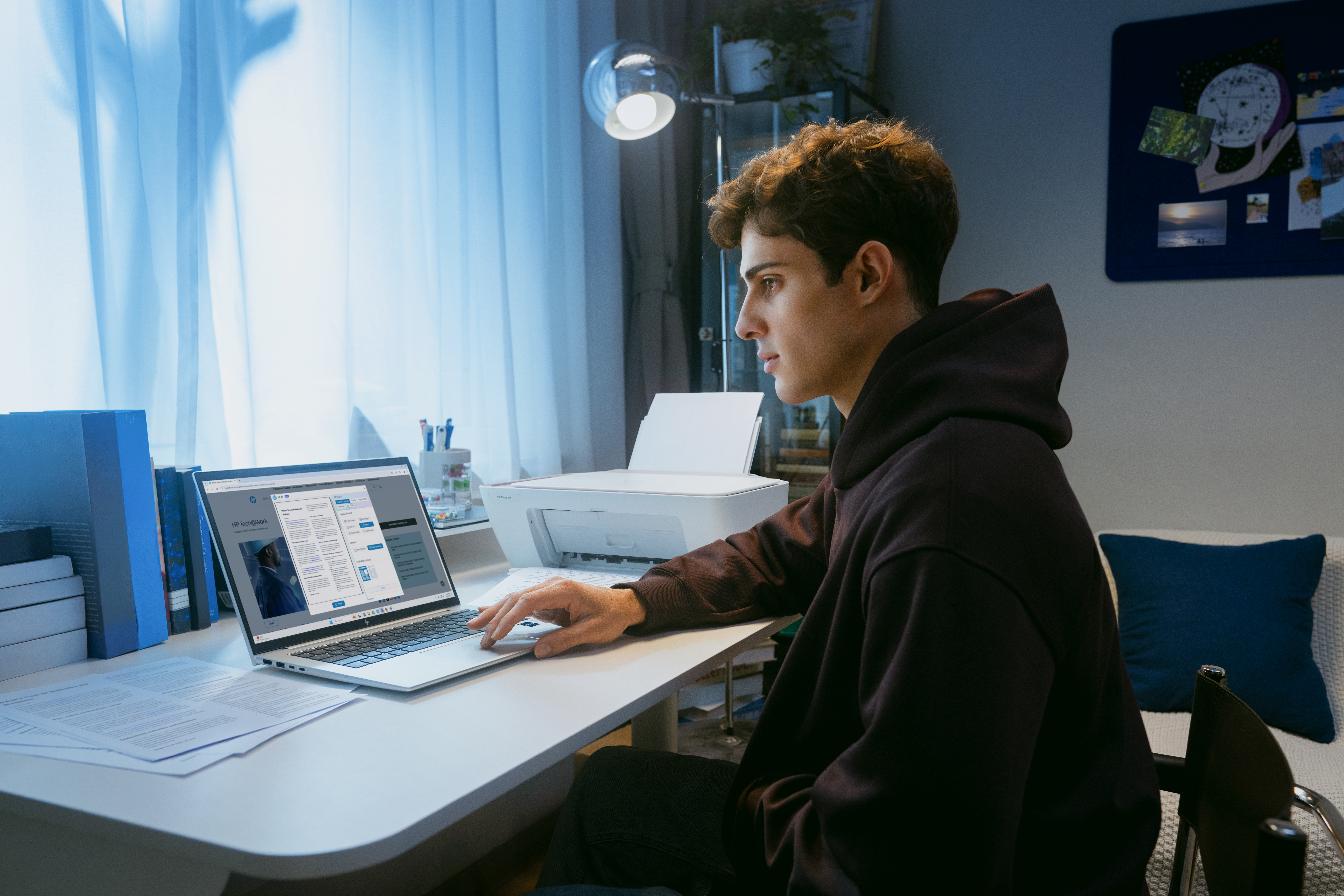 Egy fiatal barna kapucnis pulóveres férfi laptopon dolgozik az íróasztalánál este otthon.