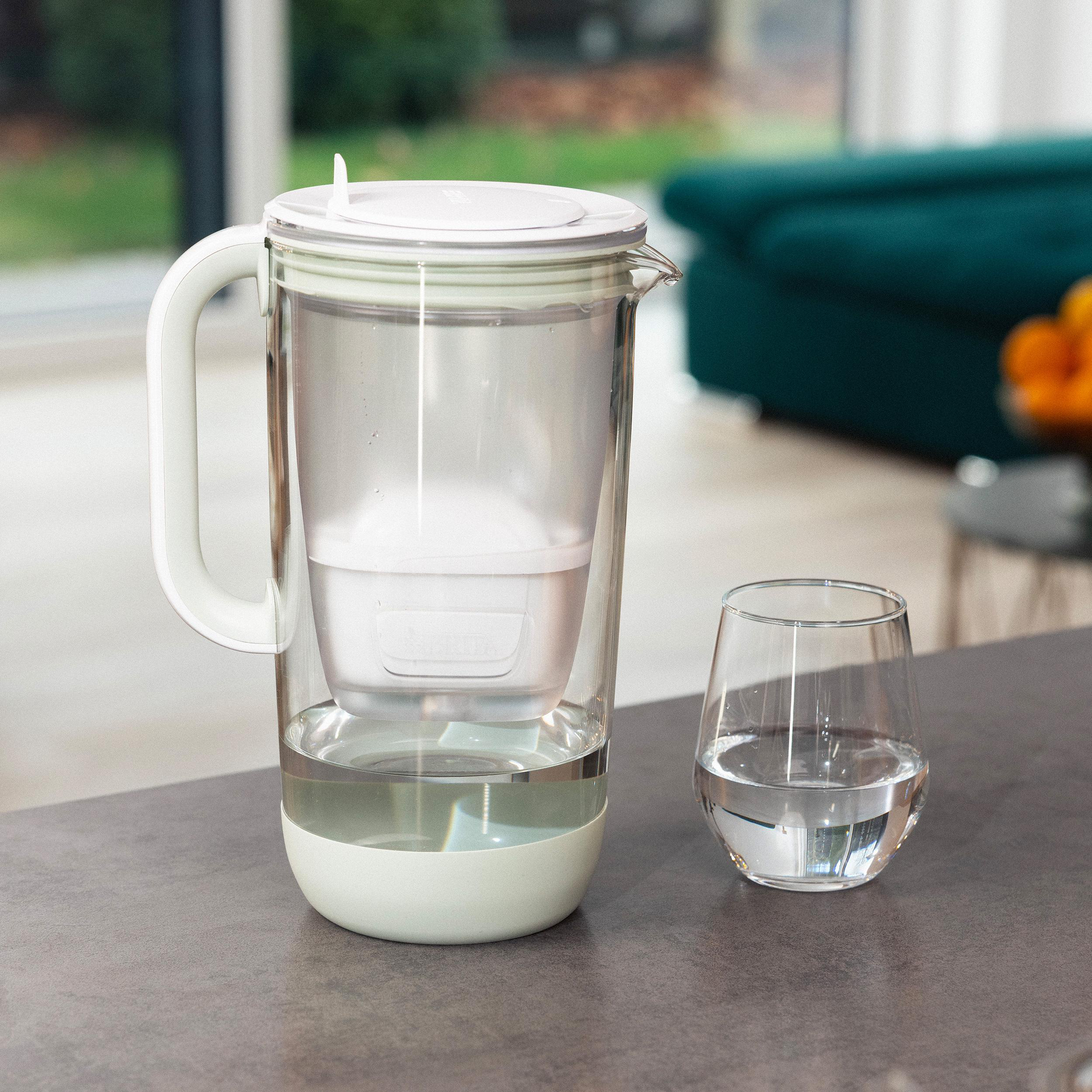 Ein klarer Wasserfilterkrug mit Griff und ein Glas Wasser auf einem grauen Tisch.