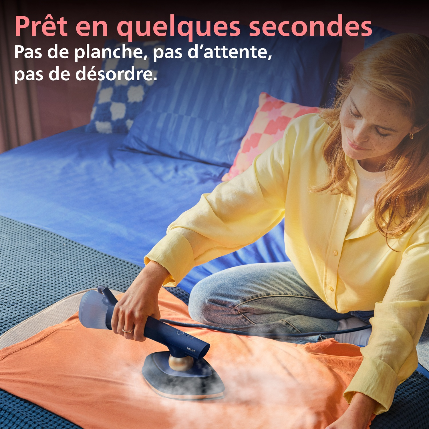 Une femme repasse une chemise orange avec un défroisseur à main sur un lit.