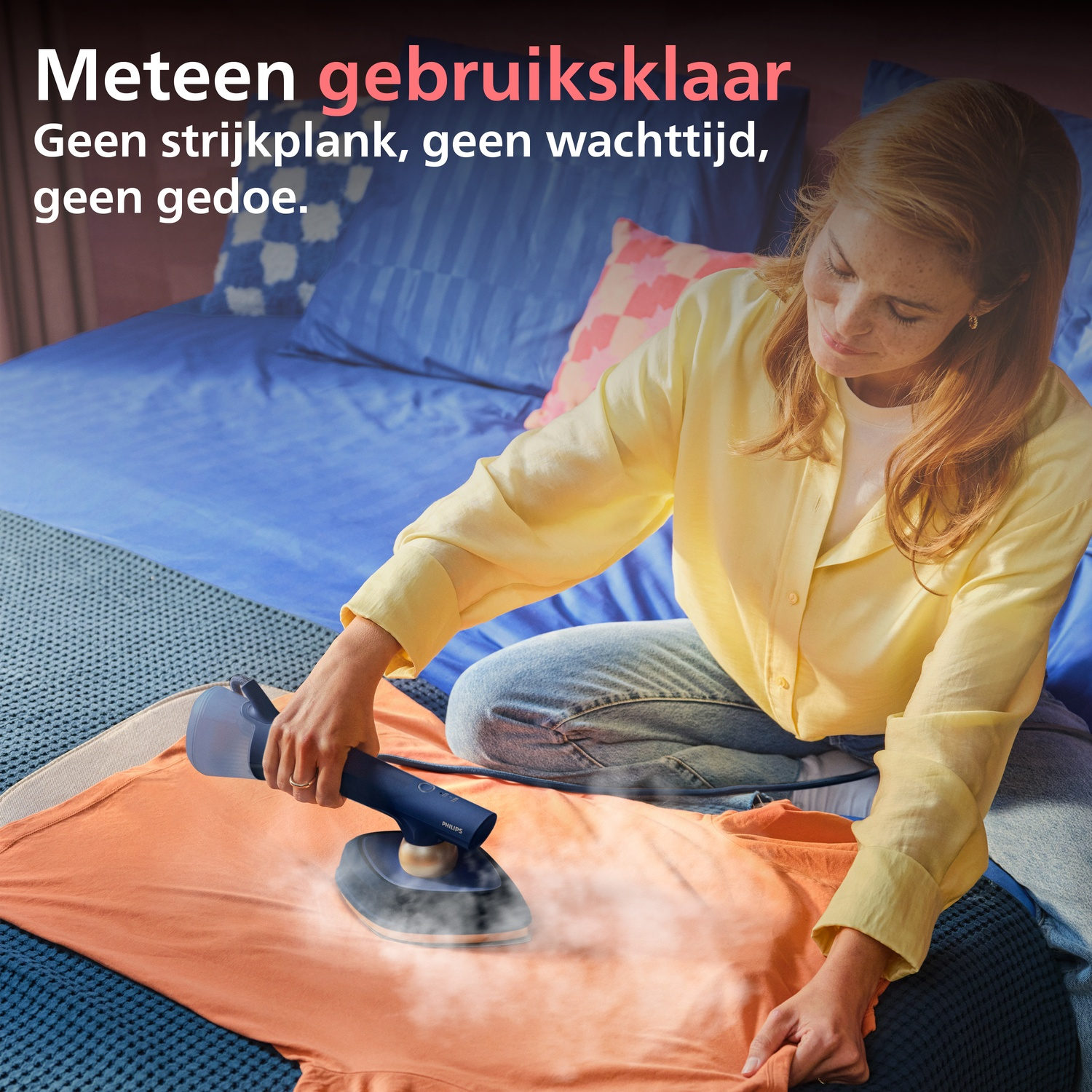 A woman irons an orange t-shirt on a bed with a handheld garment steamer.