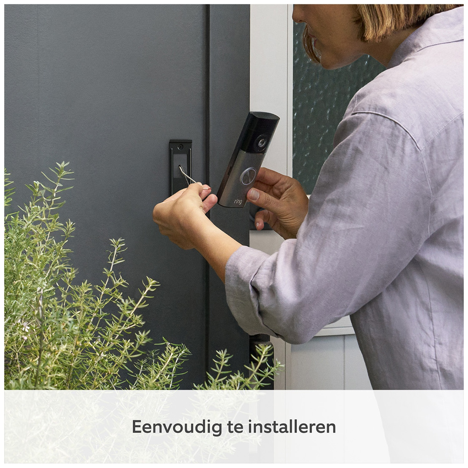Een vrouw monteert een deurbel op een grijze deur naast een groene plant.