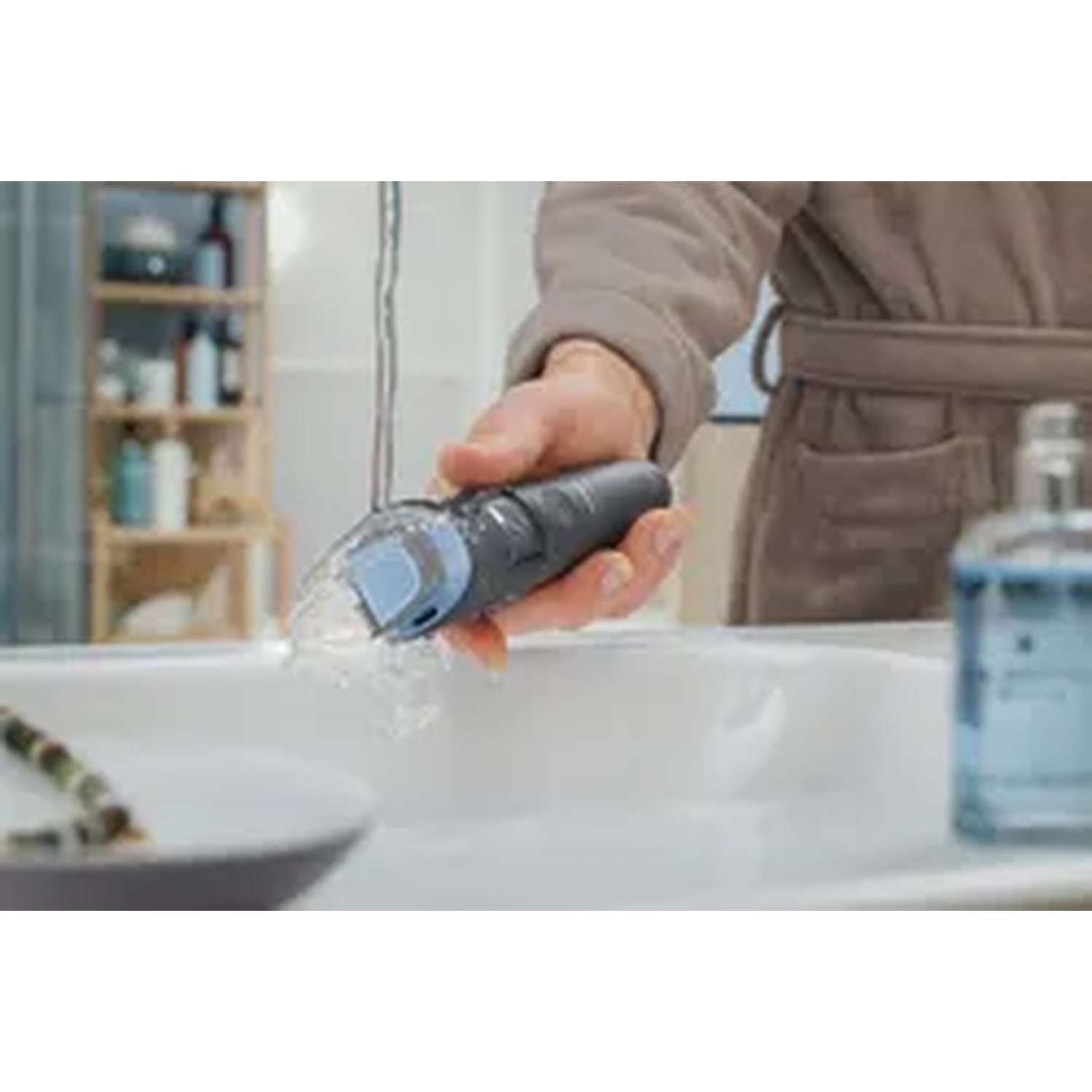 Una mano tiene un rasoio elettrico scuro sopra un lavandino, con acqua che scorre.
