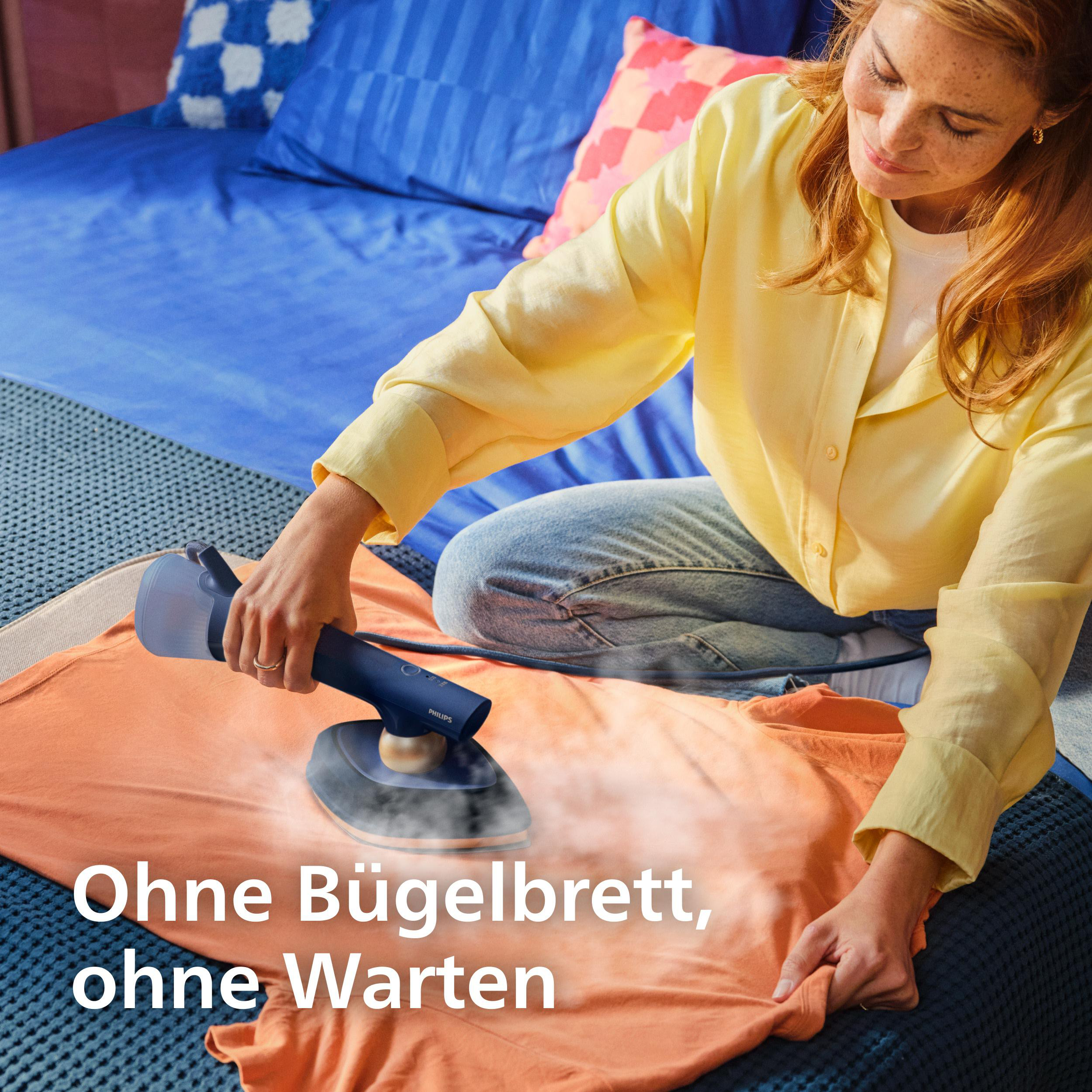 Eine Frau glättet ein orangefarbenes Hemd mit einem Handdampfer auf einem Bett.