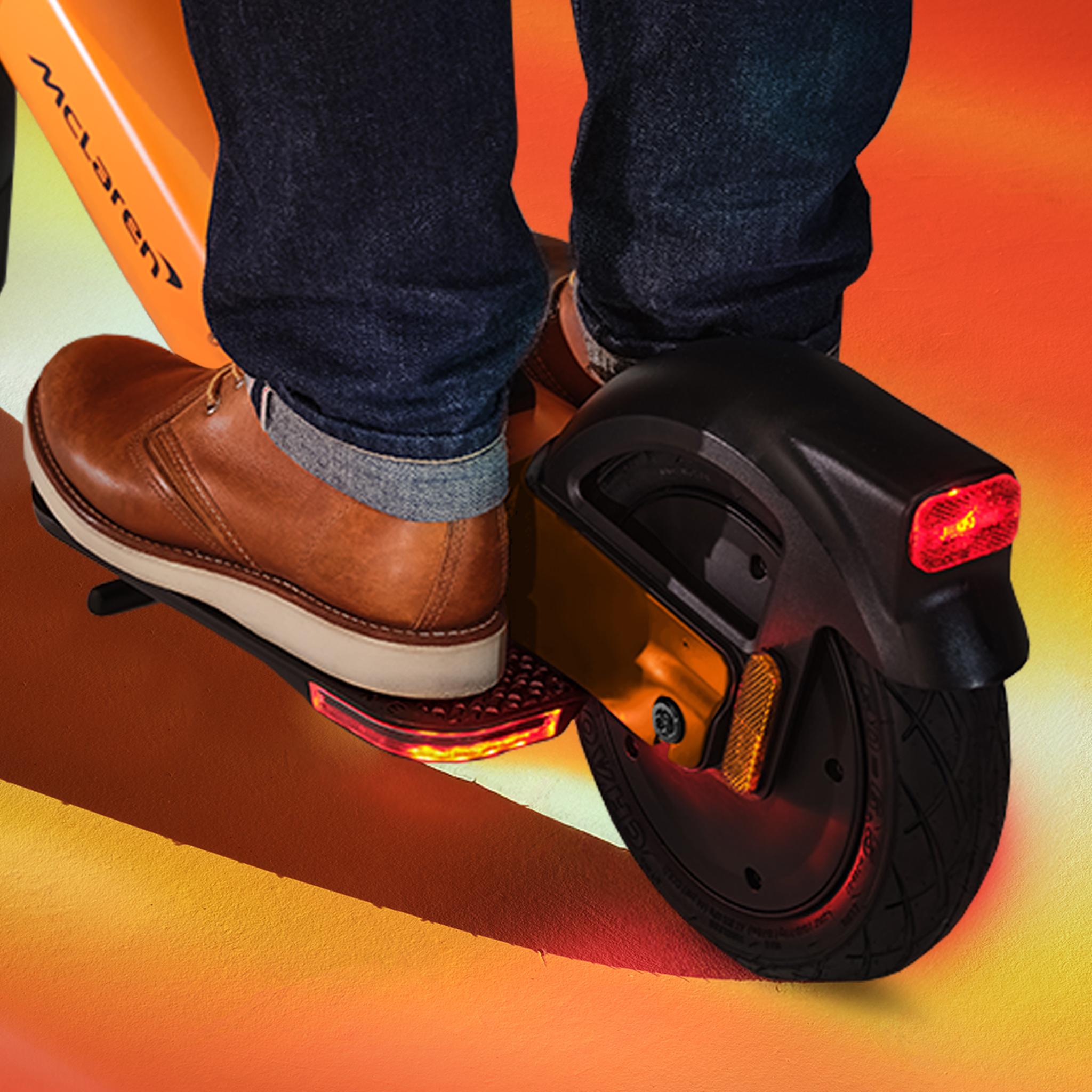 Detailaufnahme eines Mannes in Jeans und braunen Schuhen auf einem orangen Einrad mit McLaren-Logo.