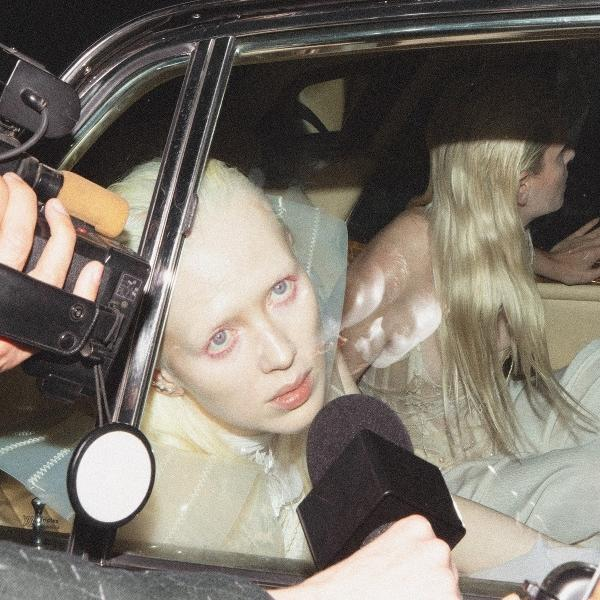 Eine Person mit heller Haut und blonden Haaren blickt aus einem Autofenster auf Kamera und Mikrofon.