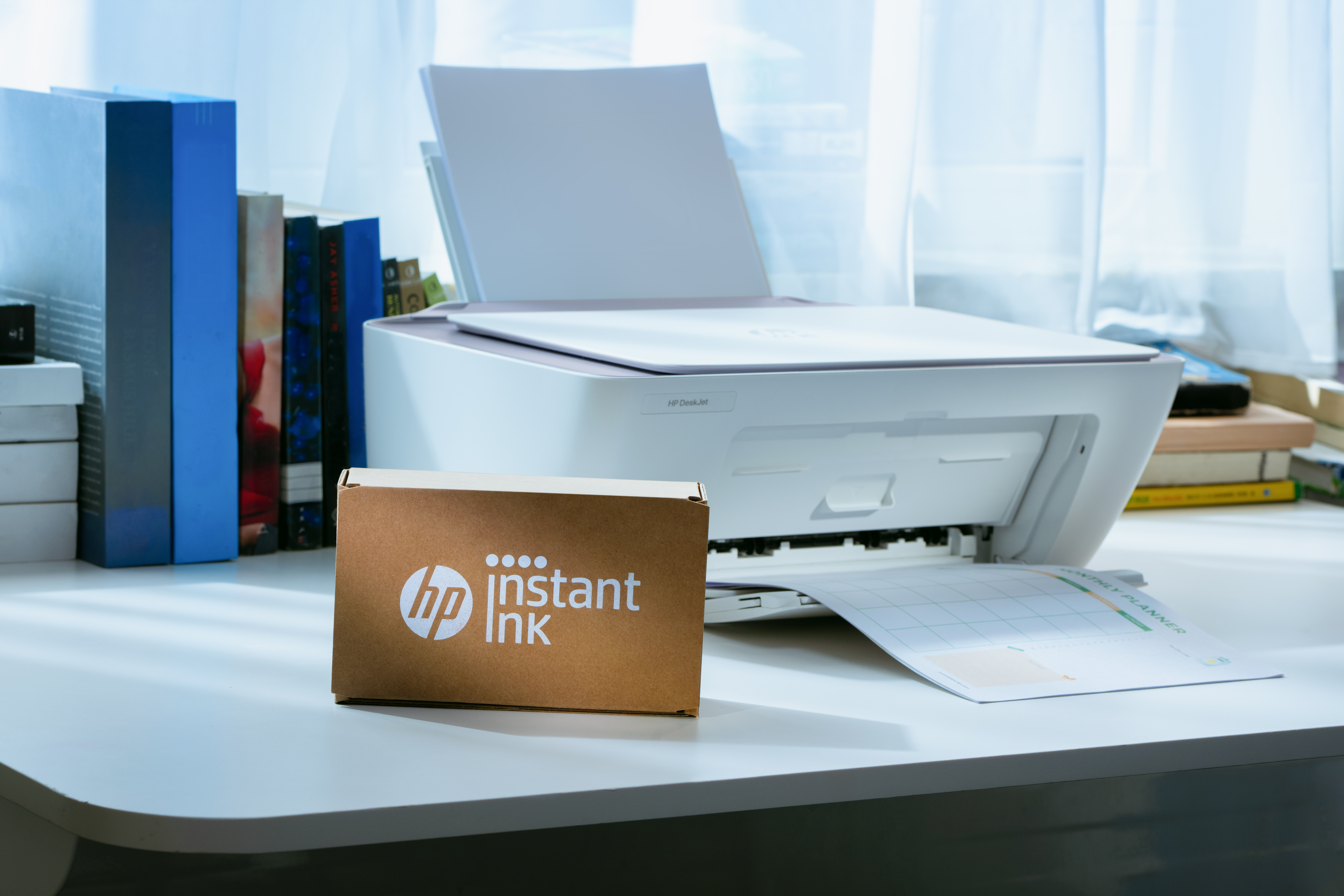 Egy HP Instant Ink doboz áll egy asztalon egy nyomtató mellett, papírral.