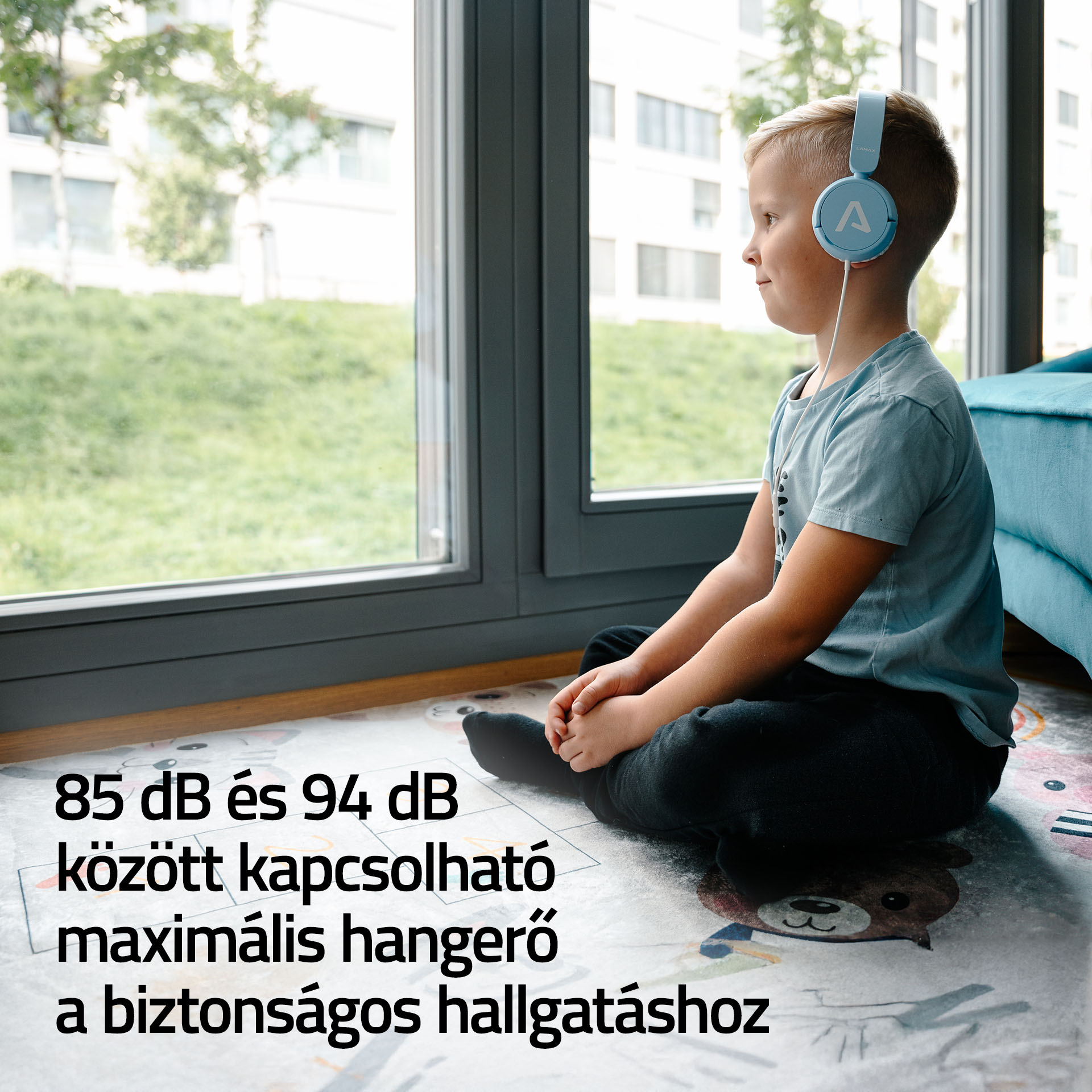 Egy fiú világoskék fejhallgatóval ül egy szőnyegen az ablaknál.