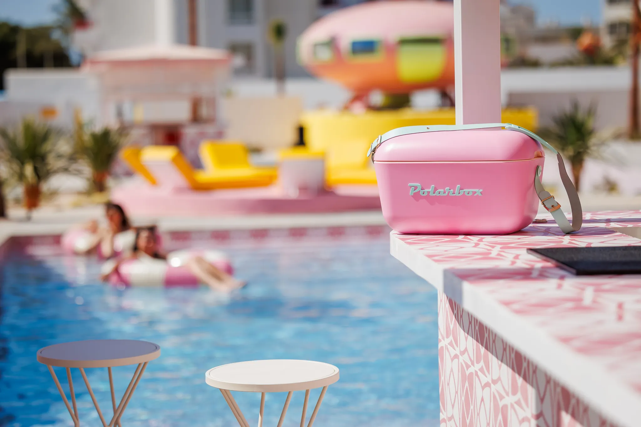 Una borsa frigo rosa è su un bancone con fantasia rosa vicino a una piscina con due persone.